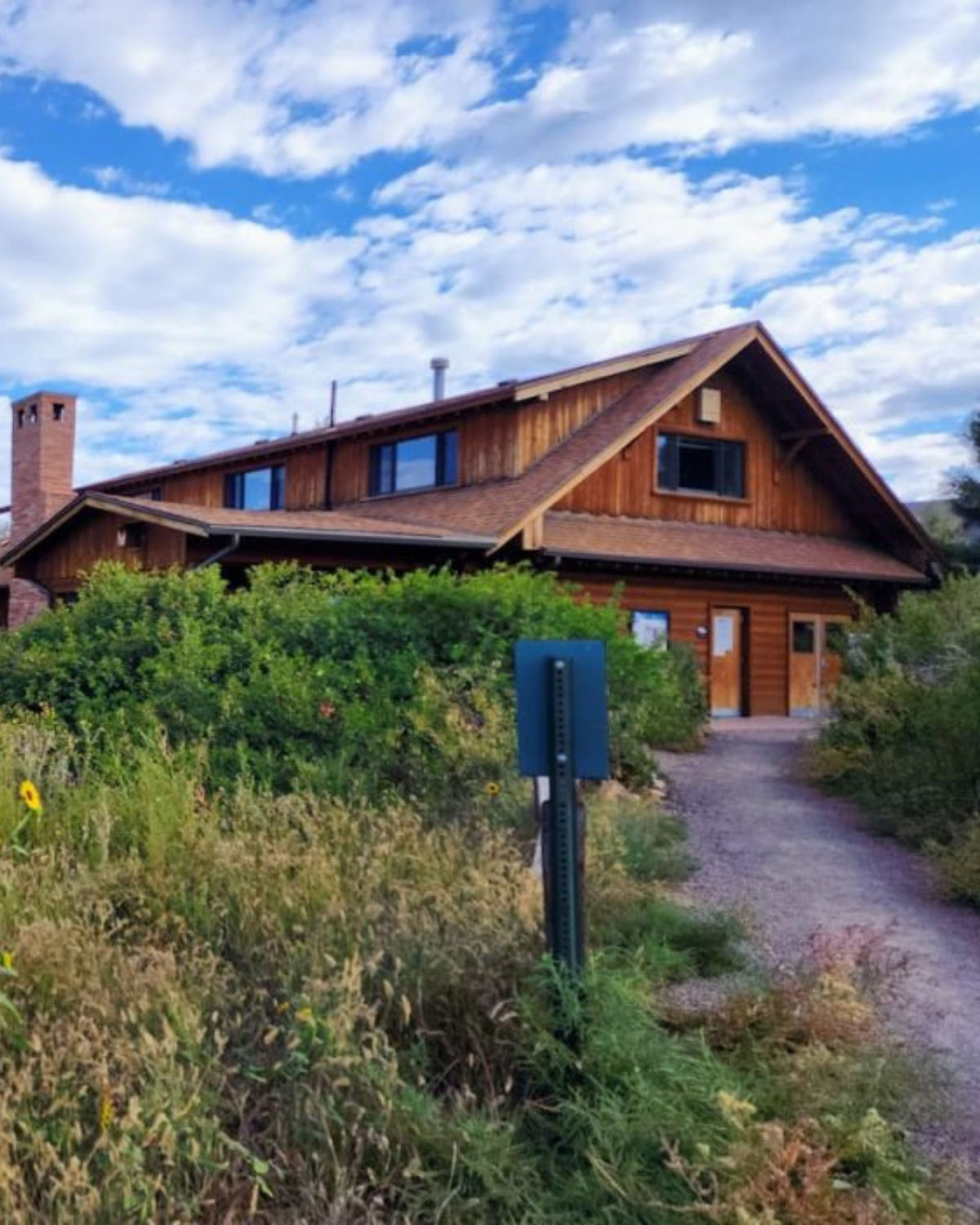 Wooden lodge-style building at South Platte Park and Carson Nature Center in Littleton, Colorado, partially hidden by tall grasses and shrubs, with a gravel path leading to the entrance under a bright, partly cloudy sky.
