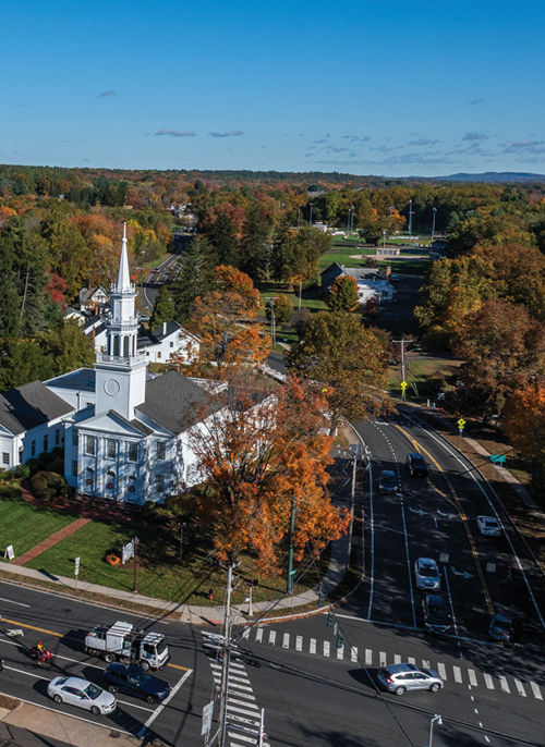 Aerial autumn view of Avon, Connecticut, featuring a historic white church with a tall steeple, tree-lined roads, colorful fall foliage, and a scenic suburban landscape stretching across the Farmington Valley beneath a clear blue sky.