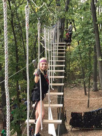 Person wearing a safety harness navigating an elevated wooden rope bridge and obstacle course suspended between tall trees at an outdoor adventure park in Bloomfield, Connecticut. The wooded setting features rope ladders, suspended platforms, and climbing obstacles.