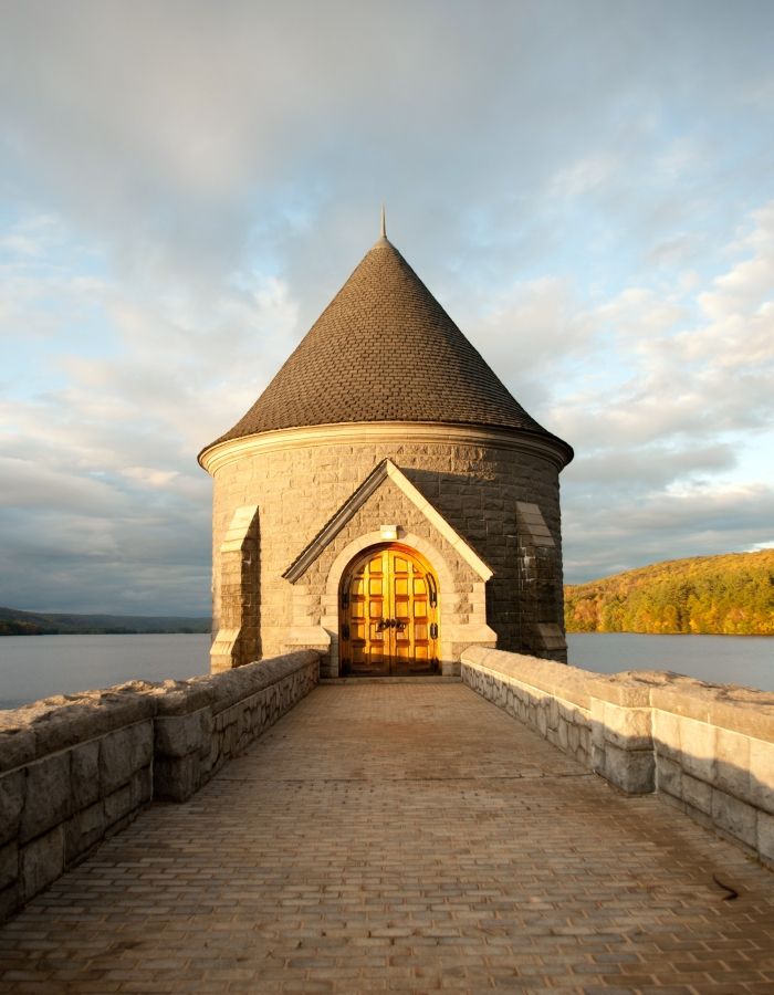 Historic stone gatehouse with a steep conical roof, arched wooden doorway, and paved stone walkway extending over a reservoir in Connecticut. Calm water, wooded hills, and golden autumn foliage surround the structure in warm late-afternoon sunlight.