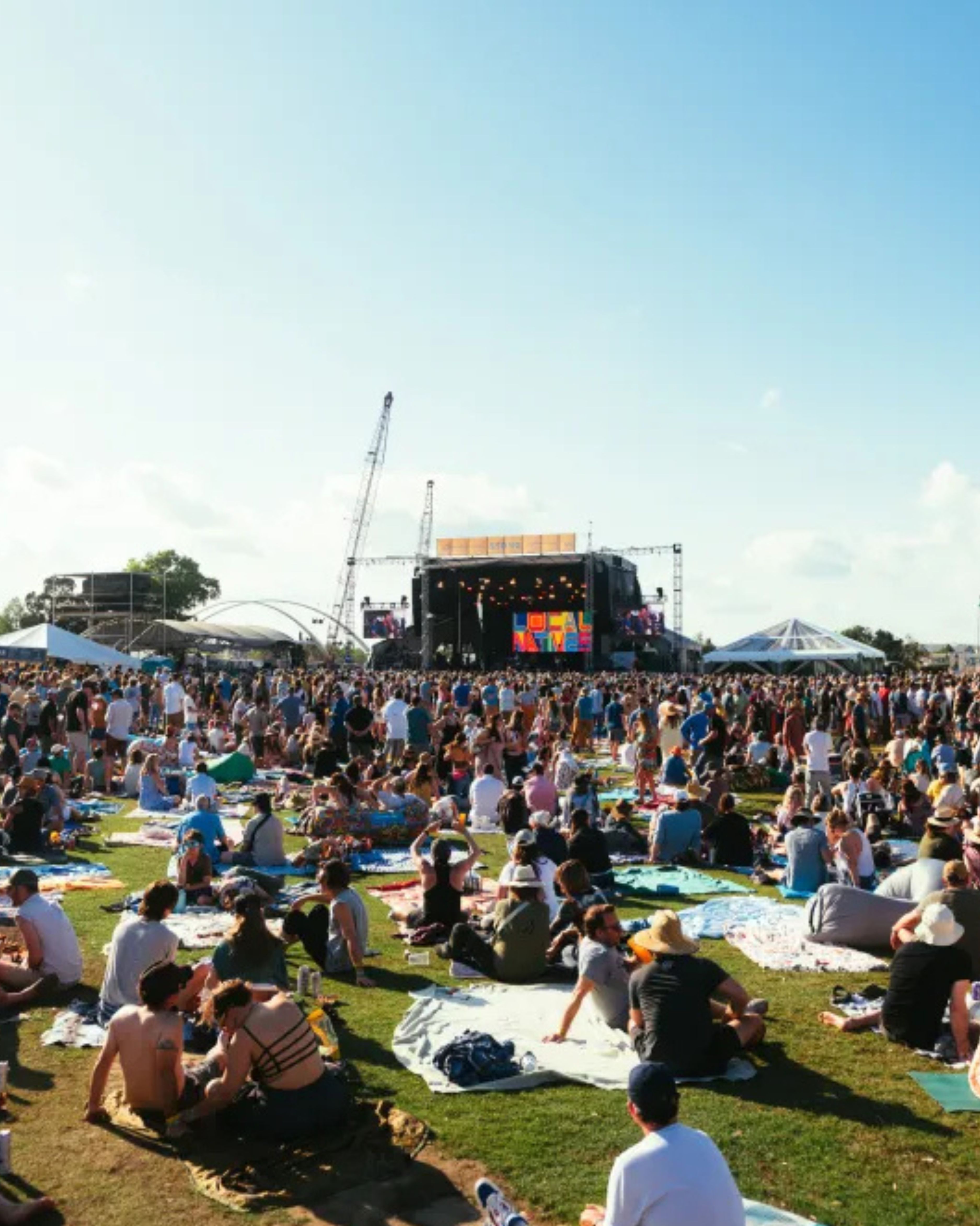 A large outdoor crowd gathered on the lawn at Seaside Park in Bridgeport, Connecticut, during a summer concert and festival. Attendees relax on blankets and lawn chairs while facing a main stage, creating a lively community atmosphere beneath clear blue skies.