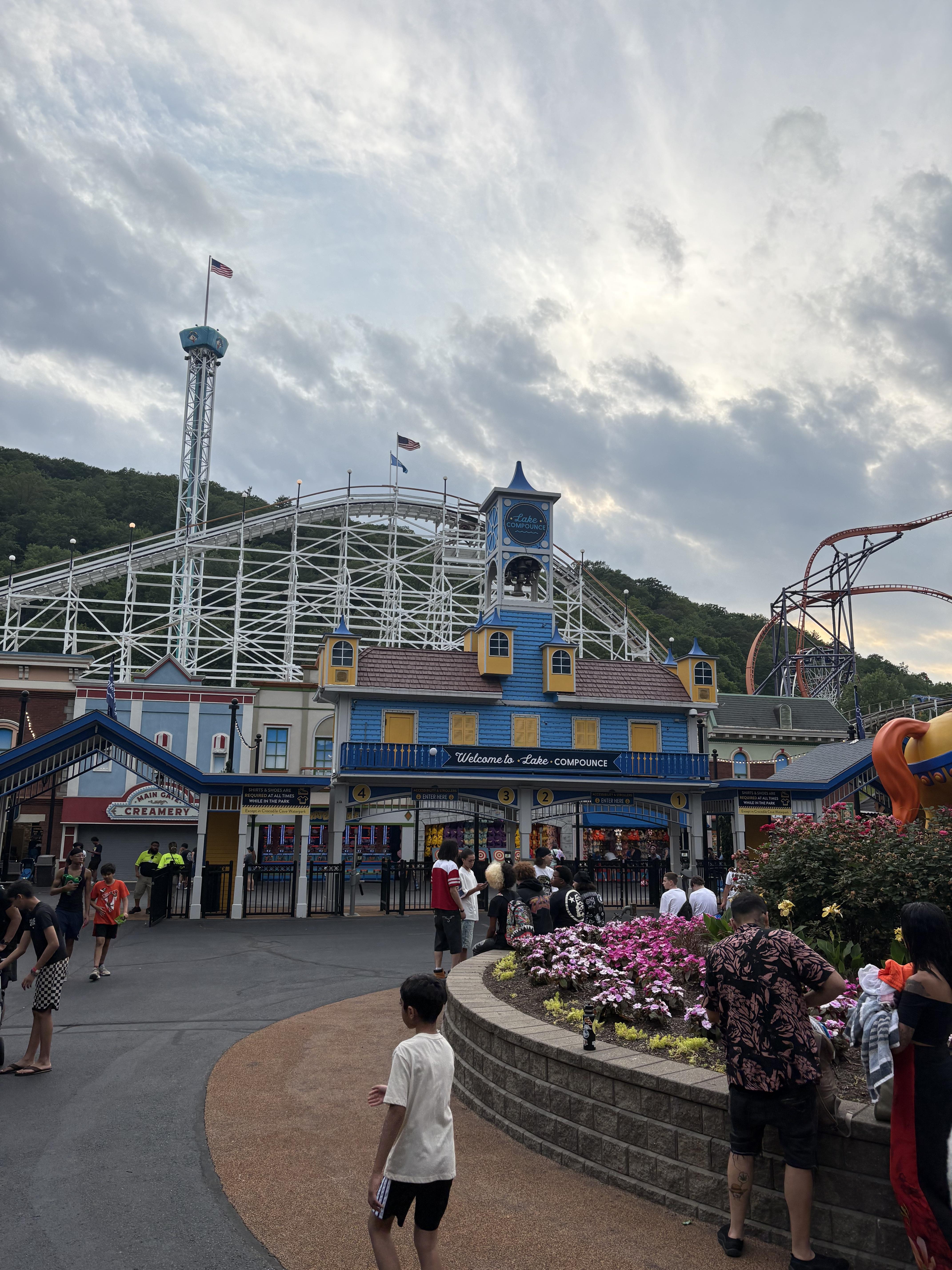 The entrance to Lake Compounce in Bristol, Connecticut, featuring the historic amusement park gateway, colorful gardens, roller coasters, and families enjoying a summer day.