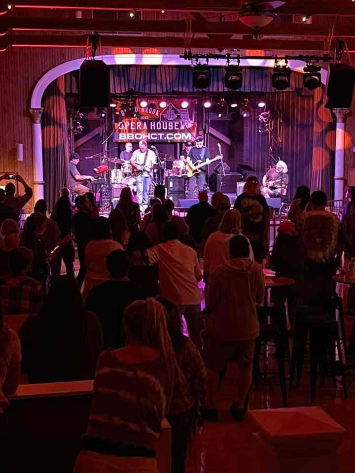 Live band performing onstage at Broad Brook Opera House in Connecticut, with musicians playing guitars, drums, and keyboard under colorful stage lighting as an audience watches.