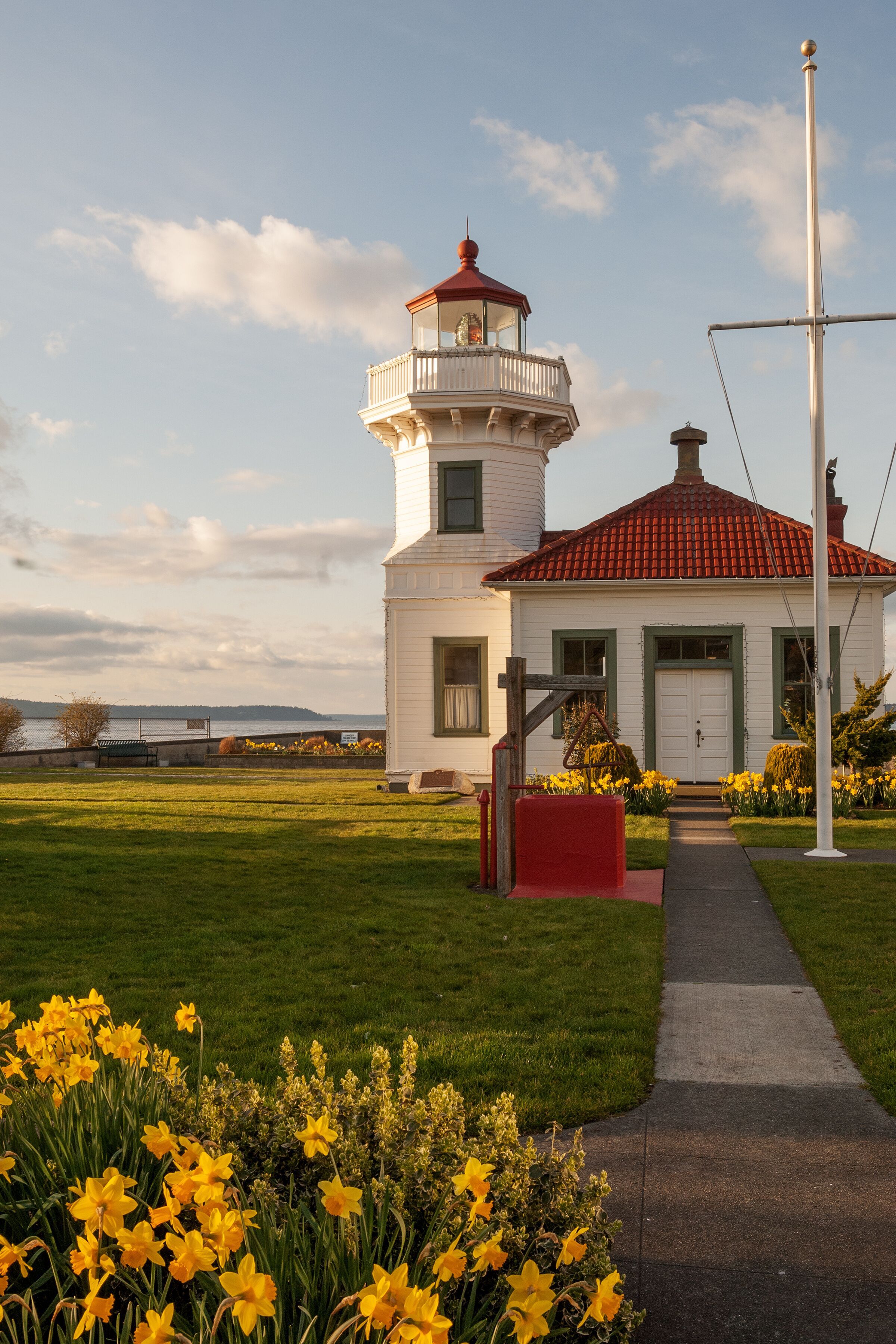 Historic white lighthouse with a red roof and lantern tower overlooking the Connecticut shoreline, surrounded by green lawns, yellow daffodils, and a waterfront walkway.