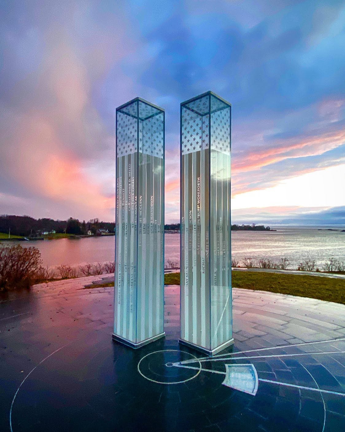 The Cos Cob 9/11 Memorial in Greenwich, Connecticut, featuring two illuminated glass towers engraved with victims' names overlooking Long Island Sound beneath a colorful evening sky.