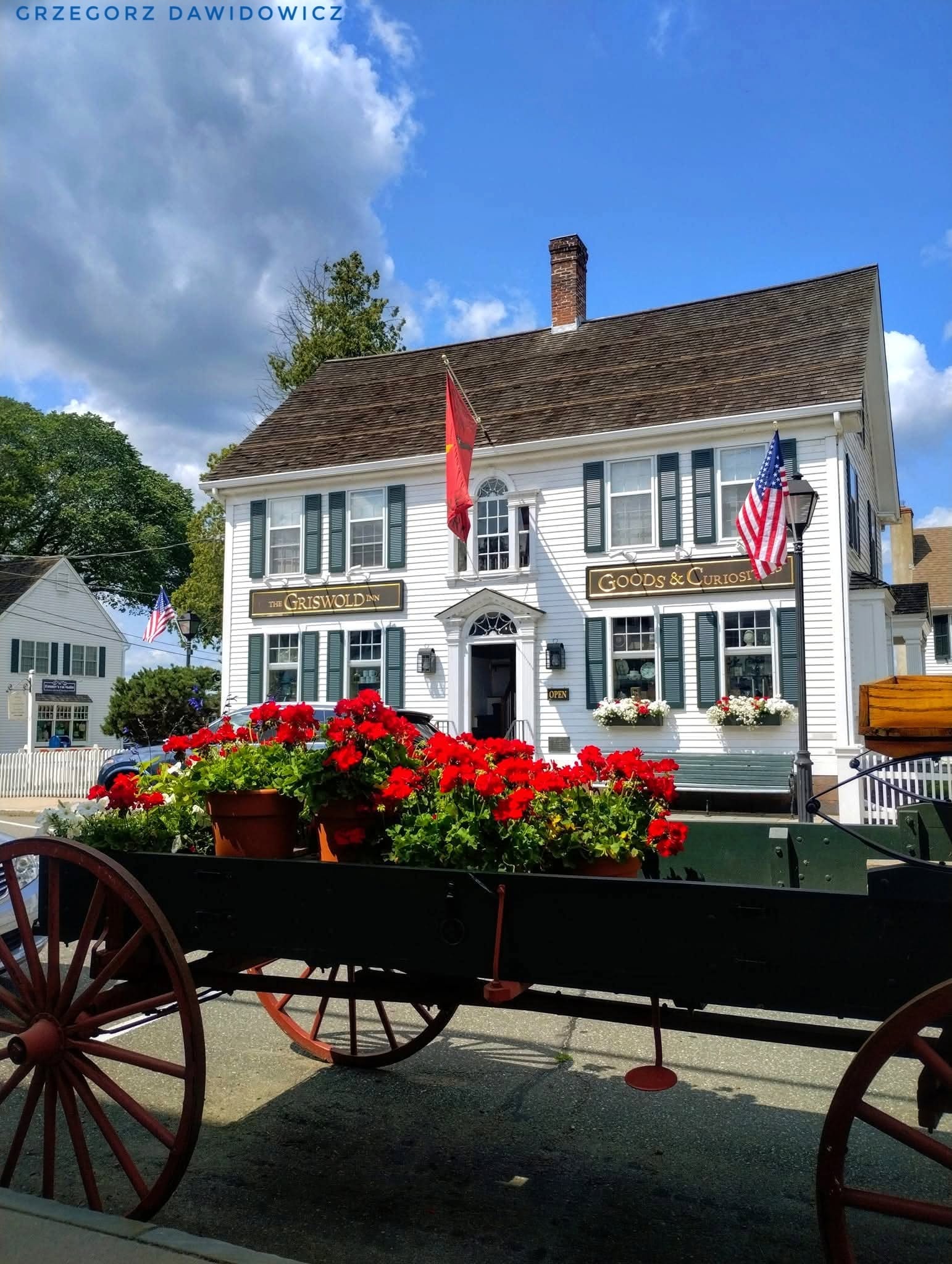Historic Griswold Inn building in Essex, Connecticut, featuring white clapboard siding, green shutters, American flags, and a wooden wagon filled with bright red flowers.