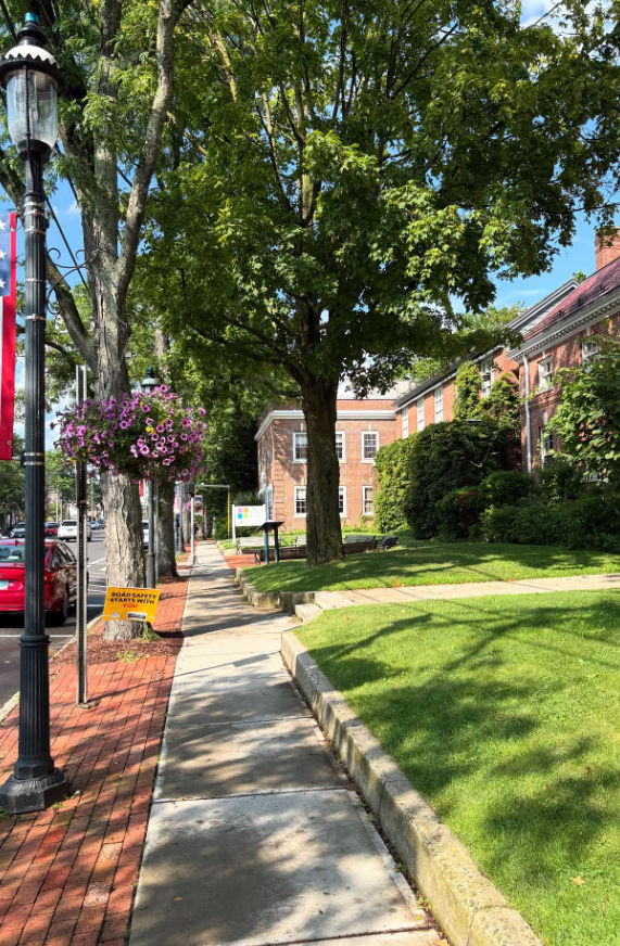 A sunny view of the Fairfield Town Green in Fairfield, Connecticut, with brick sidewalks, mature shade trees, landscaped lawns, historic buildings, and decorative hanging flower baskets lining the street.