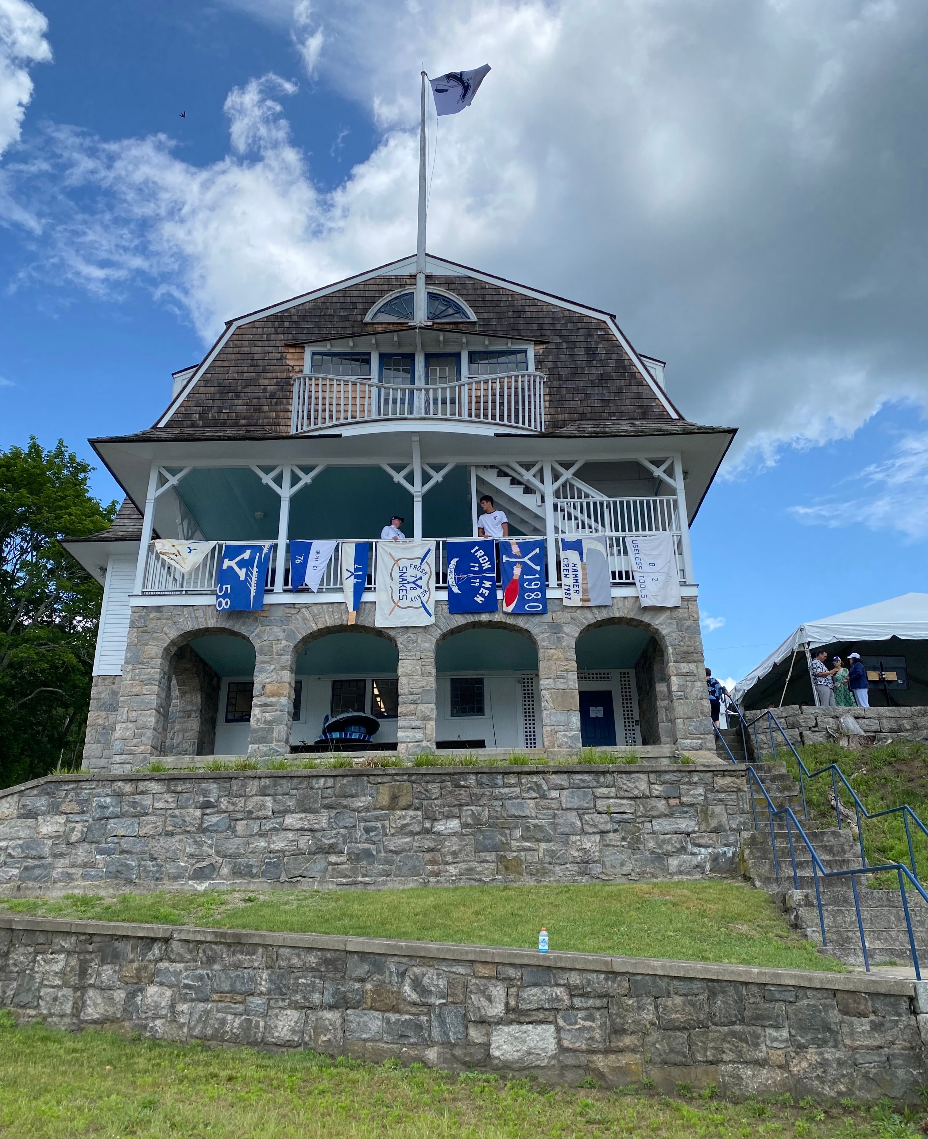 Historic waterfront rowing clubhouse associated with the Yale University crew tradition in Gales Ferry, CT, featuring a weathered shingle roof, white wooden balcony, stone archways, and rowing banners displayed along the building's exterior.