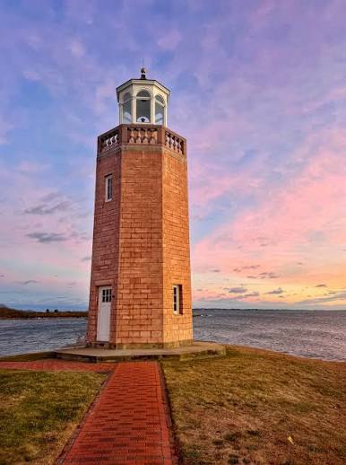 Historic brick lighthouse tower with a white lantern room overlooking Long Island Sound near Groton, CT, featuring a red brick pathway, grassy waterfront grounds, and a colorful pink and purple evening sky. The exact lighthouse is not confirmed.