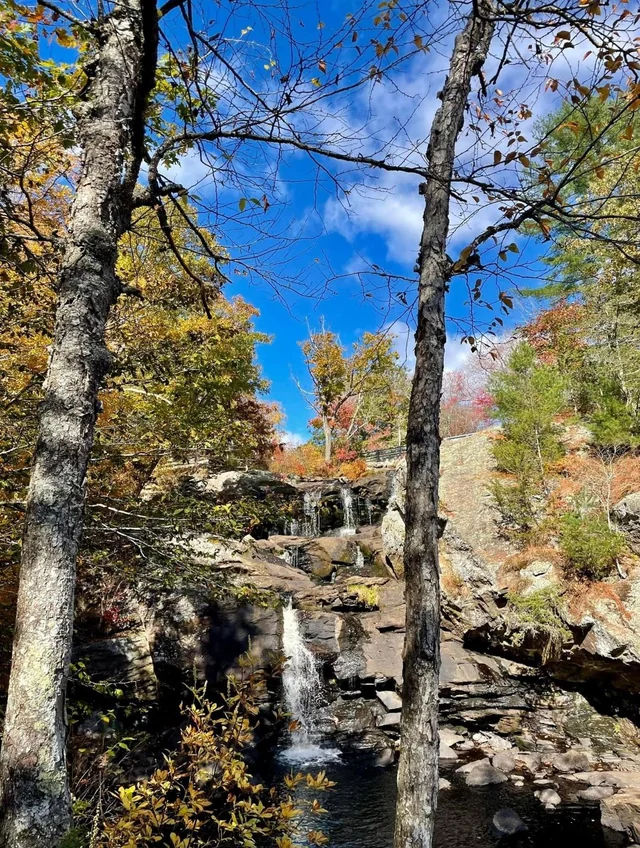 Natural woodland waterfall near Haddam, CT, featuring cascading water flowing over layered rock formations into a shallow pool, surrounded by mature trees, rugged stone ledges, and colorful early autumn foliage.