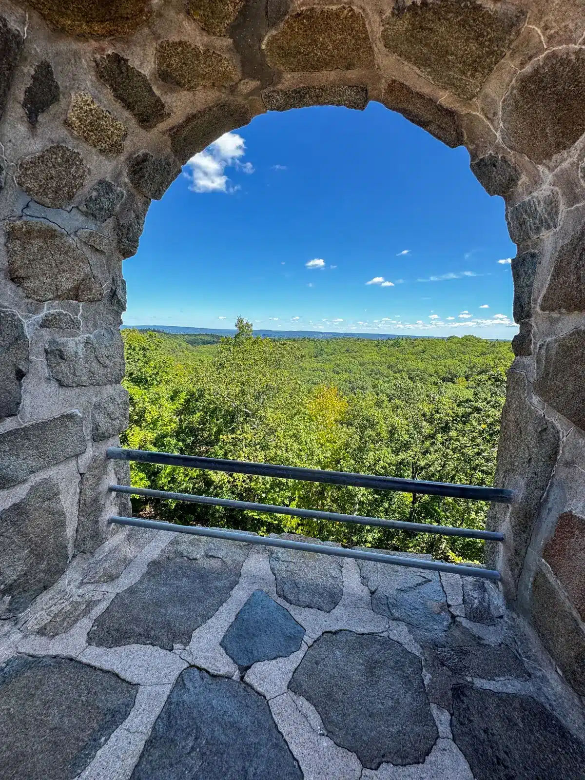 View from the stone observation tower at Sleeping Giant State Park in Hamden, Connecticut, overlooking a vast forest canopy through a historic stone archway on a clear day.