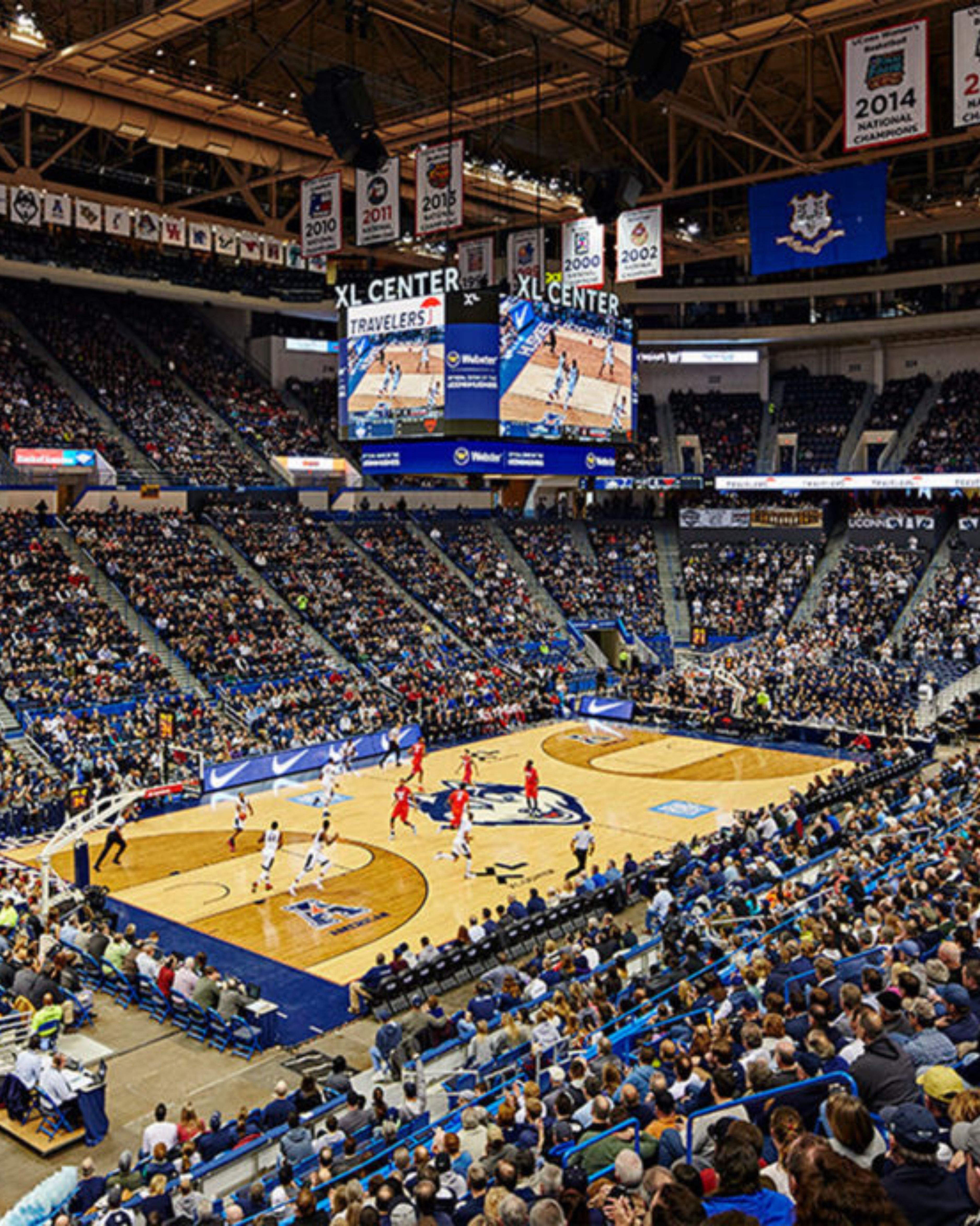 An indoor view of PeoplesBank Arena in Hartford, Connecticut, featuring championship banners hanging from the rafters, a basketball court setup below, and a spacious seating bowl that highlights the venue’s role as a hub for major sporting events and entertainment.
