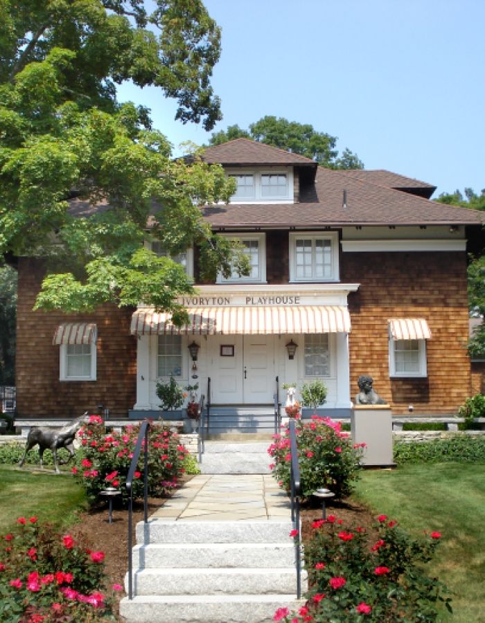 Historic Ivoryton Playhouse at 103 Main Street in Ivoryton, CT, featuring its brown shingle exterior, striped entrance awnings, white front steps, flowering rose gardens, and landscaped grounds surrounding the renowned local theater.