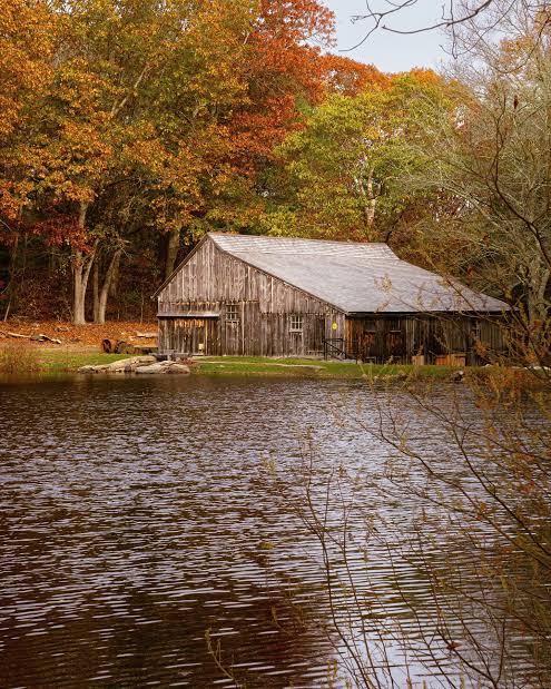 Rustic wooden waterfront building beside a quiet pond near Ledyard, CT, surrounded by mature deciduous trees displaying brilliant orange, red, and golden autumn foliage, with reflections across the water.