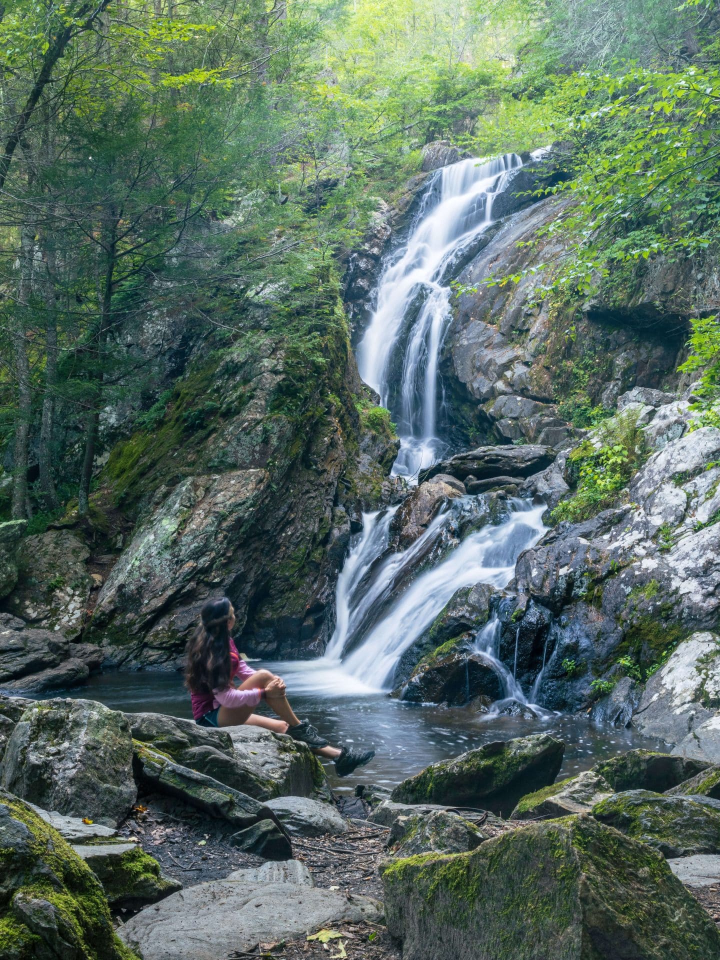 Scenic waterfall cascading over large moss-covered rocks in a wooded natural area near Marlborough, Connecticut, with a visitor sitting beside the water surrounded by lush green trees.