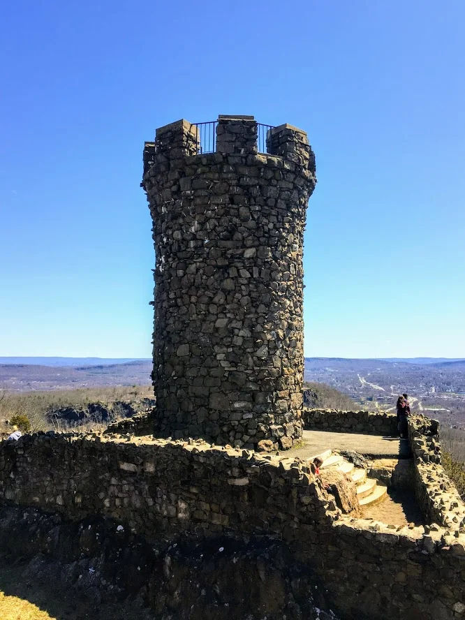 A close-up view of the historic stone Castle Craig observation tower atop East Peak in Meriden, Connecticut, surrounded by stone walls and offering sweeping views of the surrounding hills under a clear blue sky.