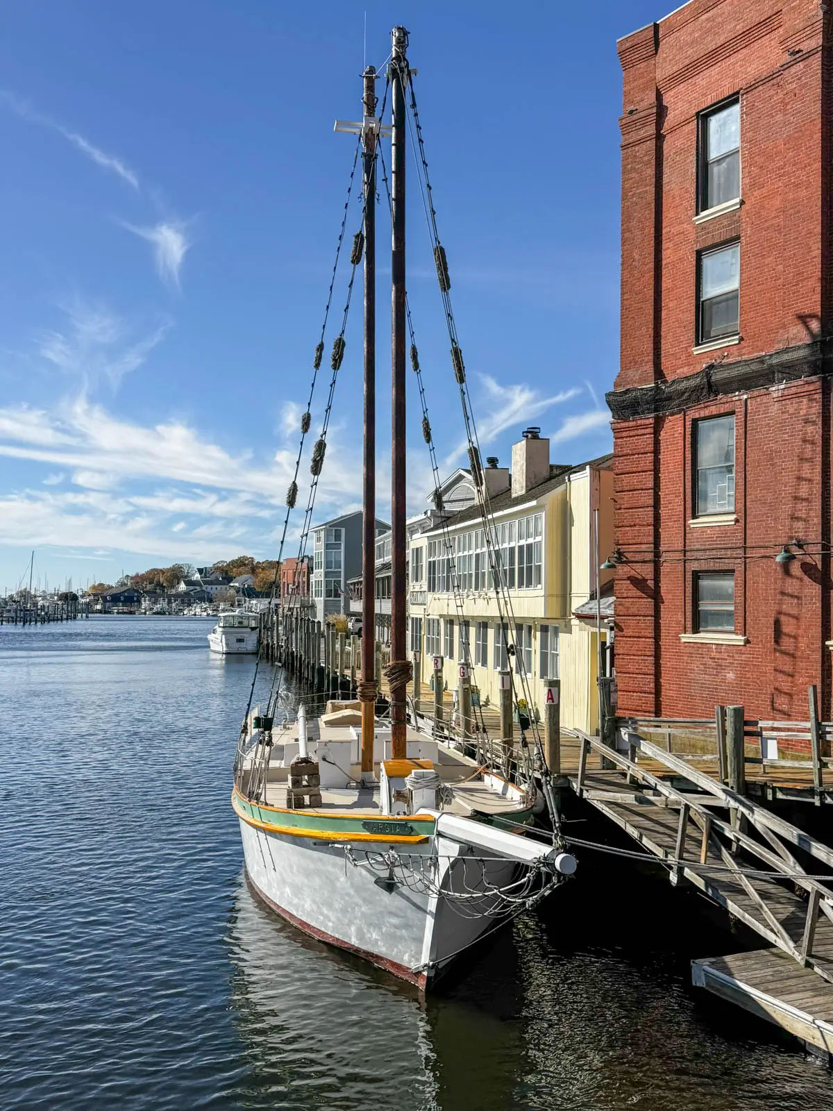 Historic sailing vessel docked along the Mystic, Connecticut, waterfront, featuring tall wooden masts, waterfront buildings, wooden docks, and calm harbor waters beneath a bright blue sky.