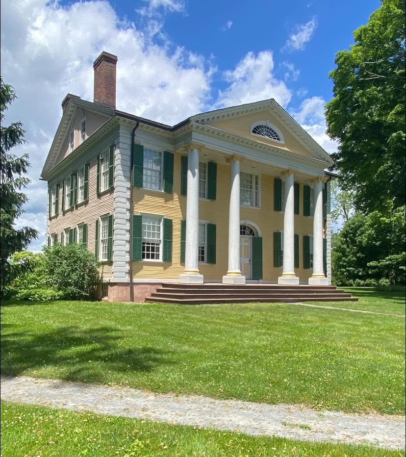 Historic colonial-style building in Old Lyme, Connecticut, featuring pale yellow siding, green shutters, tall white columns, and a manicured lawn beneath a blue summer sky.