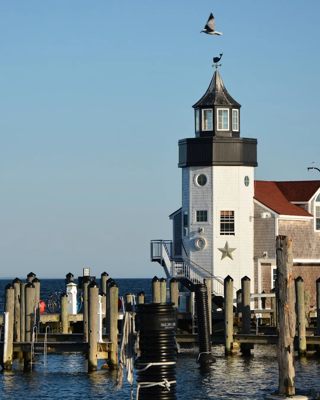 Lighthouse-style marina building at Saybrook Point Marina in Old Saybrook, CT, featuring a white shingle exterior, black cupola, decorative nautical details, wooden docks, and boat slips overlooking the Connecticut River near Long Island Sound.