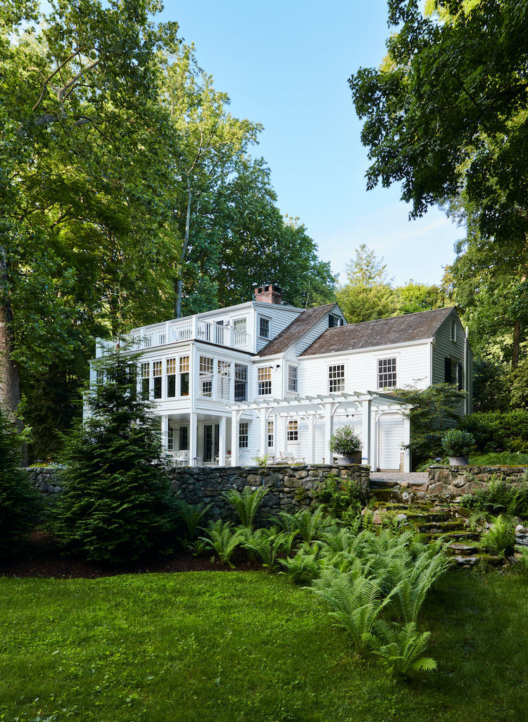 The historic Bush-Holley House in Riverside, Connecticut, a beautifully preserved white colonial home surrounded by mature trees, landscaped gardens, and a classic New England stone terrace.