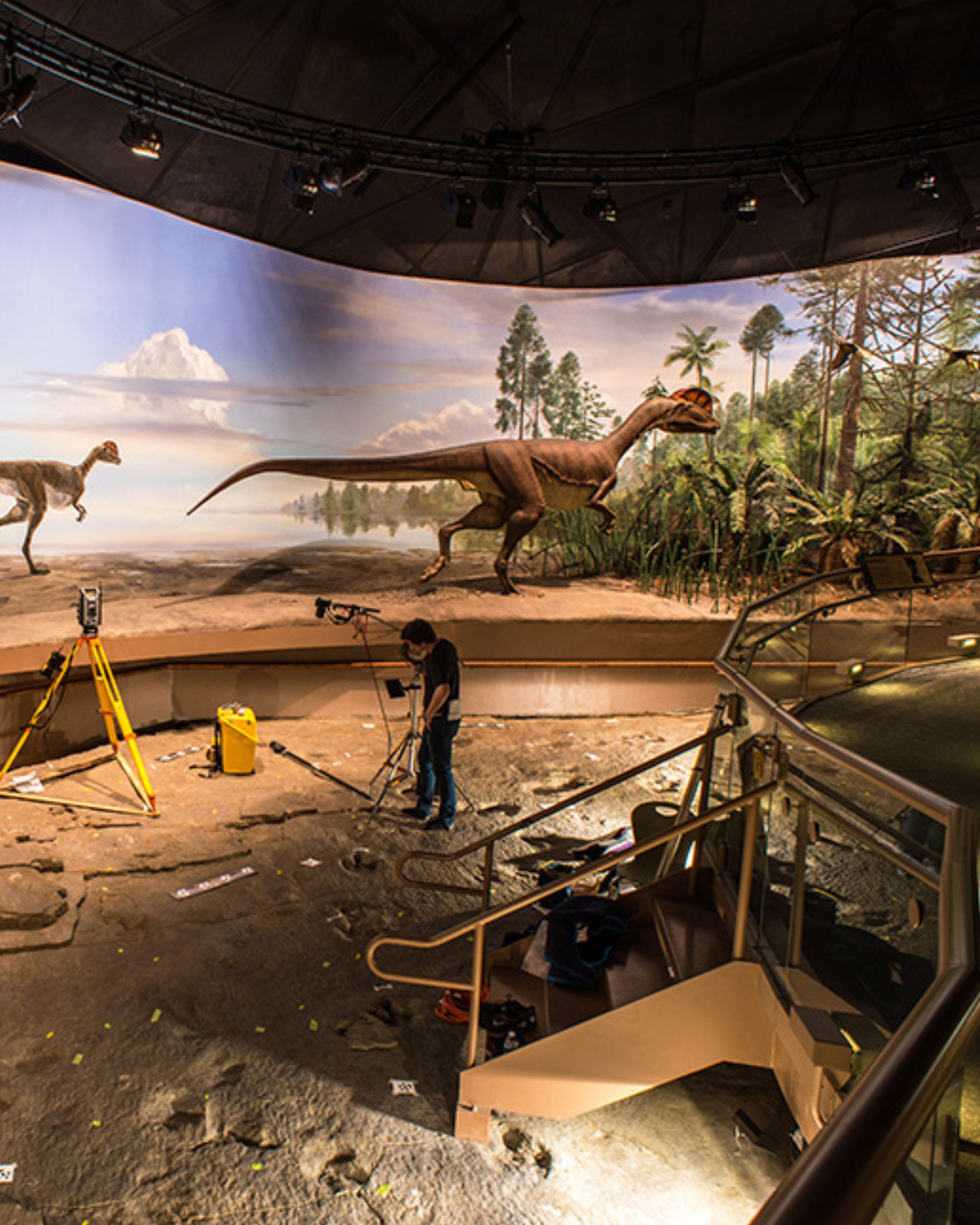Wide-angle interior view of the dinosaur exhibit hall at Dinosaur State Park in Rocky Hill, Connecticut, featuring large prehistoric dinosaur displays, excavation-style terrain, overhead theatrical lighting, stair railings, and a museum worker operating surveying and camera equipment during exhibit setup.