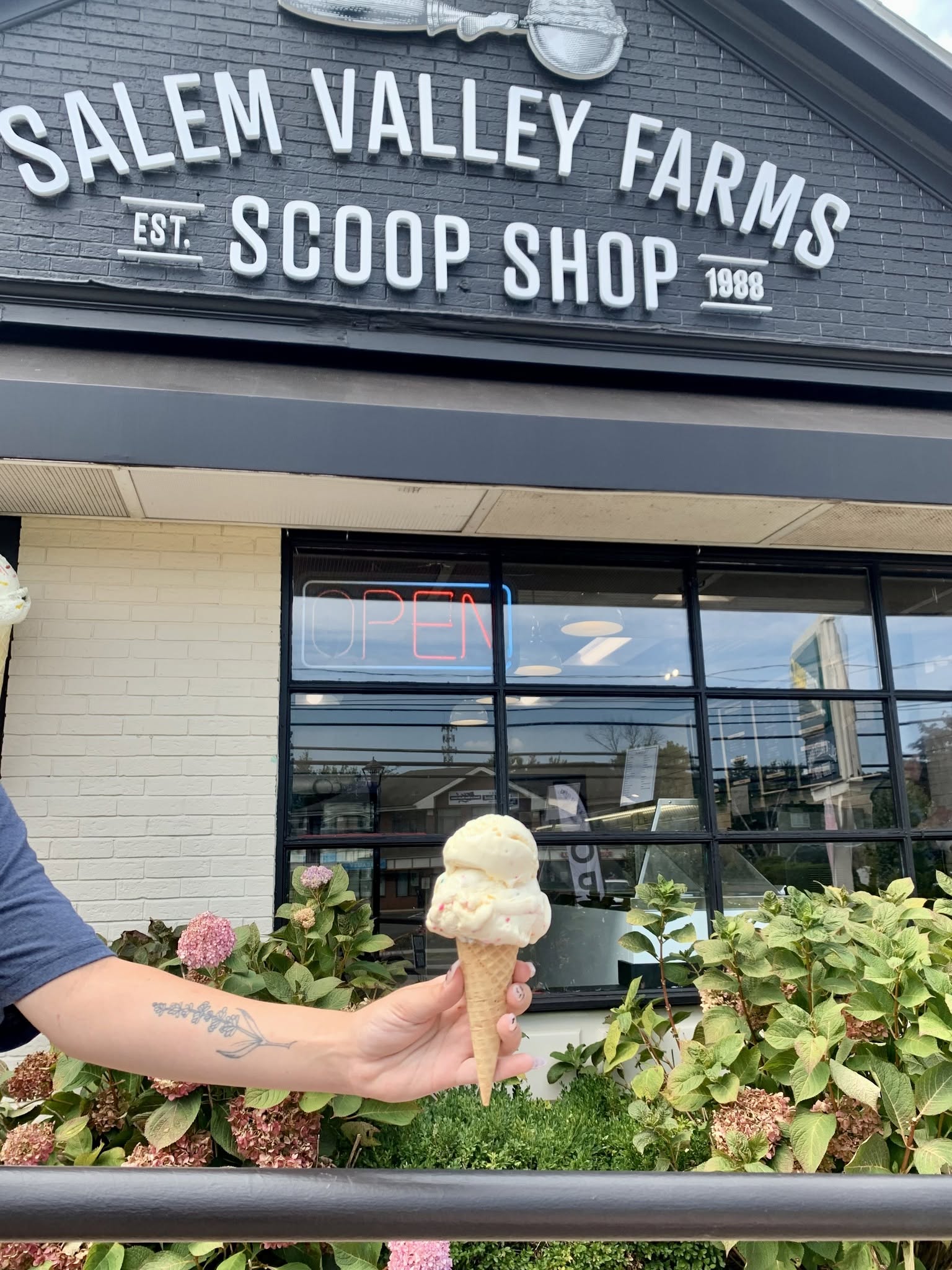 Salem Valley Farms Scoop Shop storefront in Salem, Connecticut, featuring a hand holding an ice cream cone outside the local ice cream shop, with an illuminated open sign and landscaped flowers.
