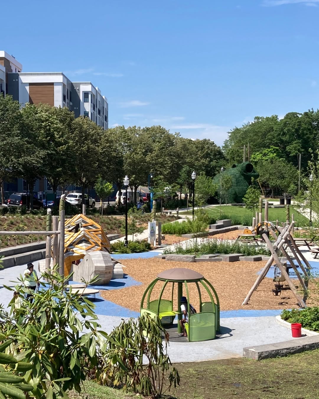 Children's playground and community park in Stamford, Connecticut, featuring nature-inspired play structures, walking paths, landscaped gardens, and open green space. Surrounded by trees and modern residential buildings, the park provides a welcoming outdoor gathering place for families.
