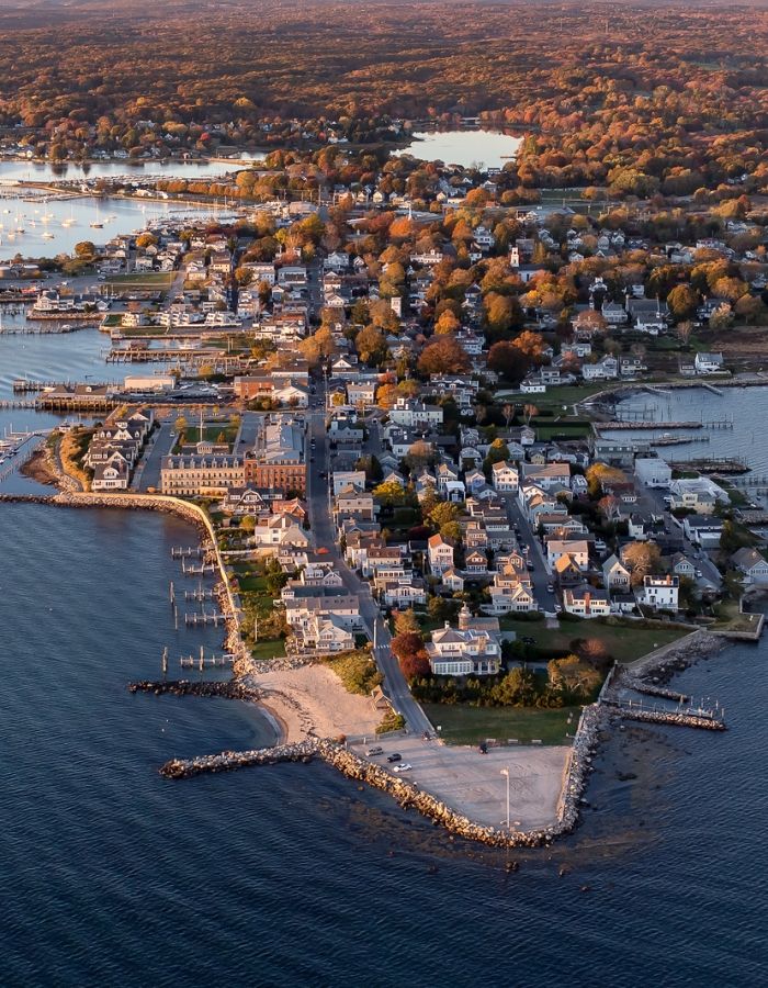 Aerial view of Stonington Borough in Stonington, CT, featuring densely clustered historic coastal homes, narrow streets, waterfront marinas, stone breakwaters, sandy shoreline, and surrounding waters of Fishers Island Sound beneath colorful autumn foliage.