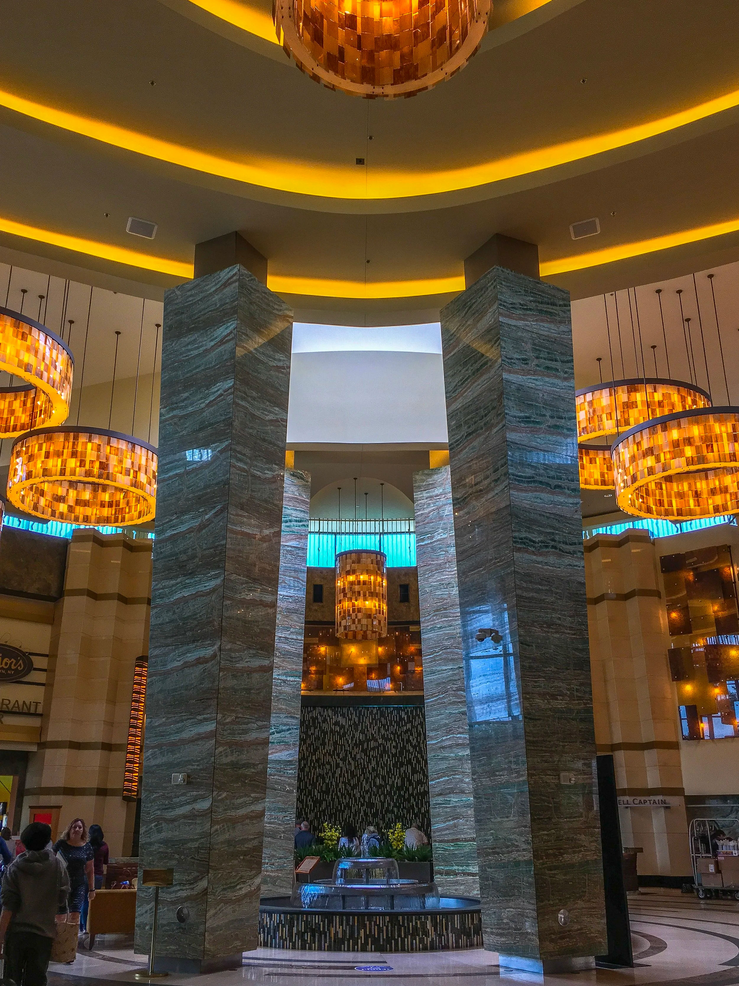 Interior of Mohegan Sun Casino and Resort in Uncasville, CT, featuring the grand Casino of the Sky architectural design, towering stone-clad columns, illuminated circular chandeliers, curved ceiling lighting, and a central decorative fountain surrounded by restaurants and entertainment spaces.
