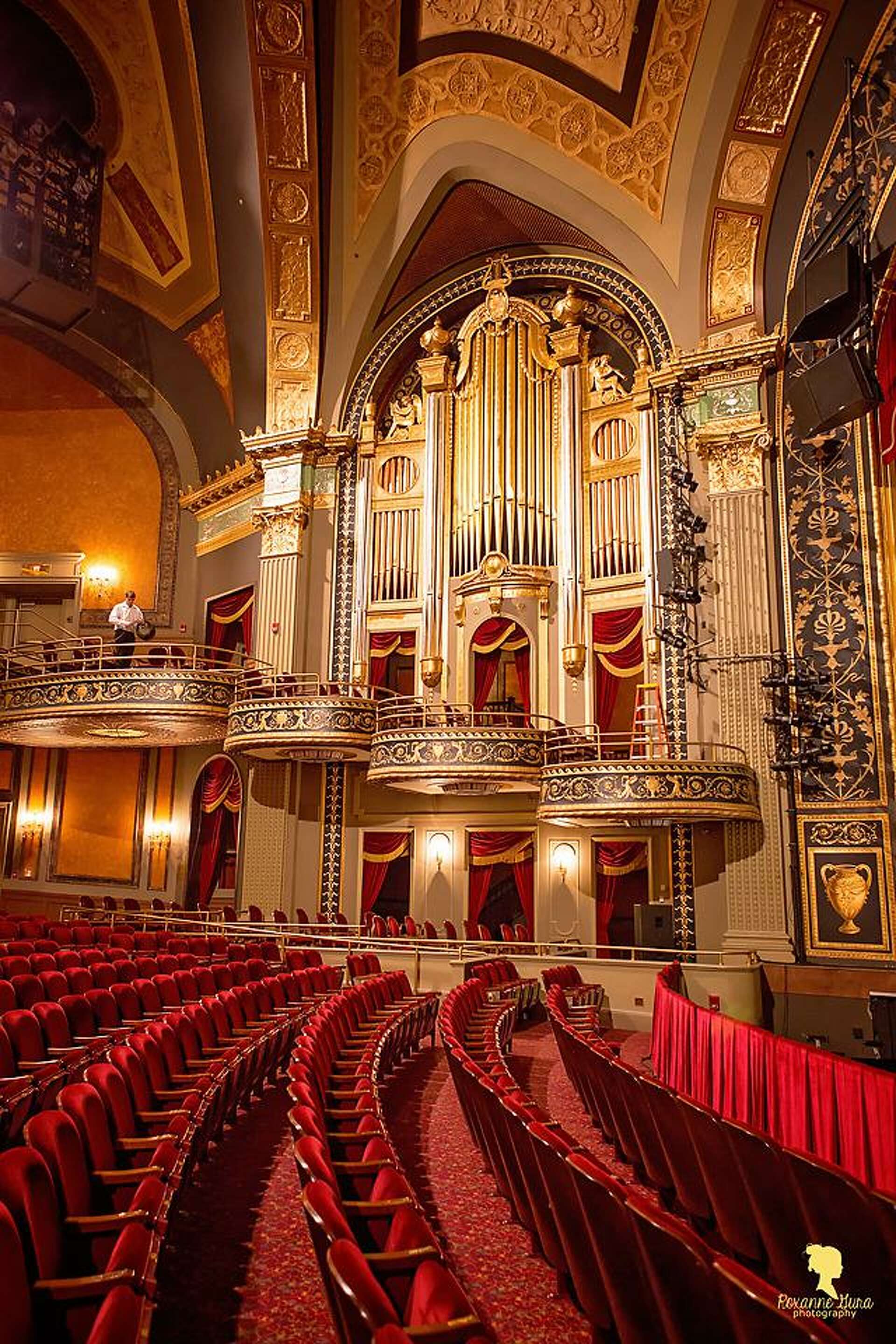 The ornate interior of the Palace Theater in Waterbury, Connecticut, showcasing rows of red velvet seats, decorative balconies, gold detailing, and a grand pipe organ. The historic performing arts venue features elegant architecture and classic theater design illuminated by warm lighting.