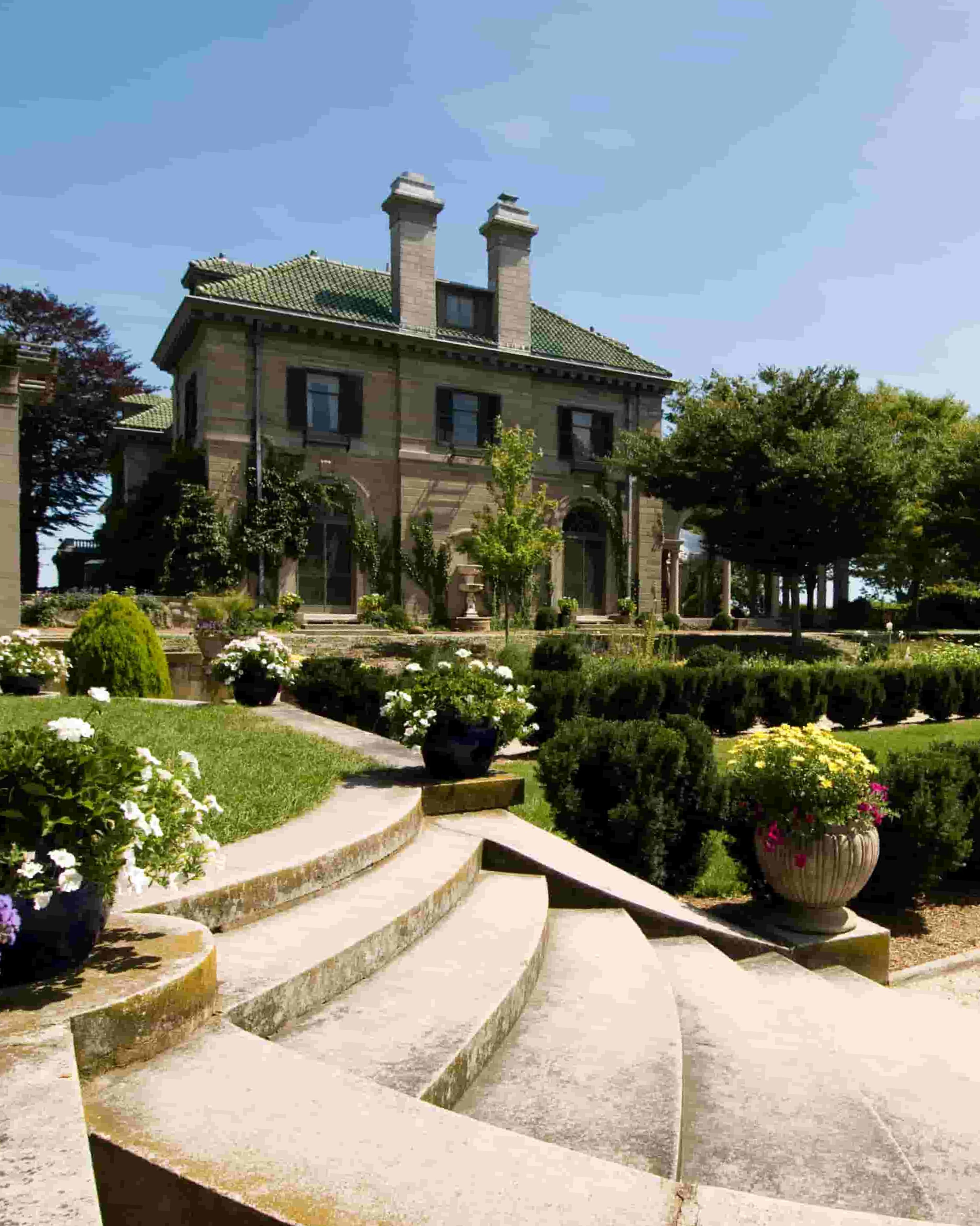 View of Harkness Memorial State Park in Waterford, Connecticut, featuring manicured formal gardens, blooming flowers, trimmed hedges, and pergola-style architectural elements on a sunny day.
