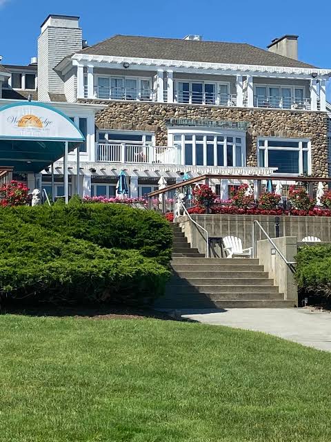 Exterior view of the Water's Edge Resort & Spa in Westbrook, CT, featuring its stone-and-white-trim waterfront architecture, outdoor terraces, landscaped flower gardens, central staircase, and manicured green lawn overlooking Long Island Sound.
