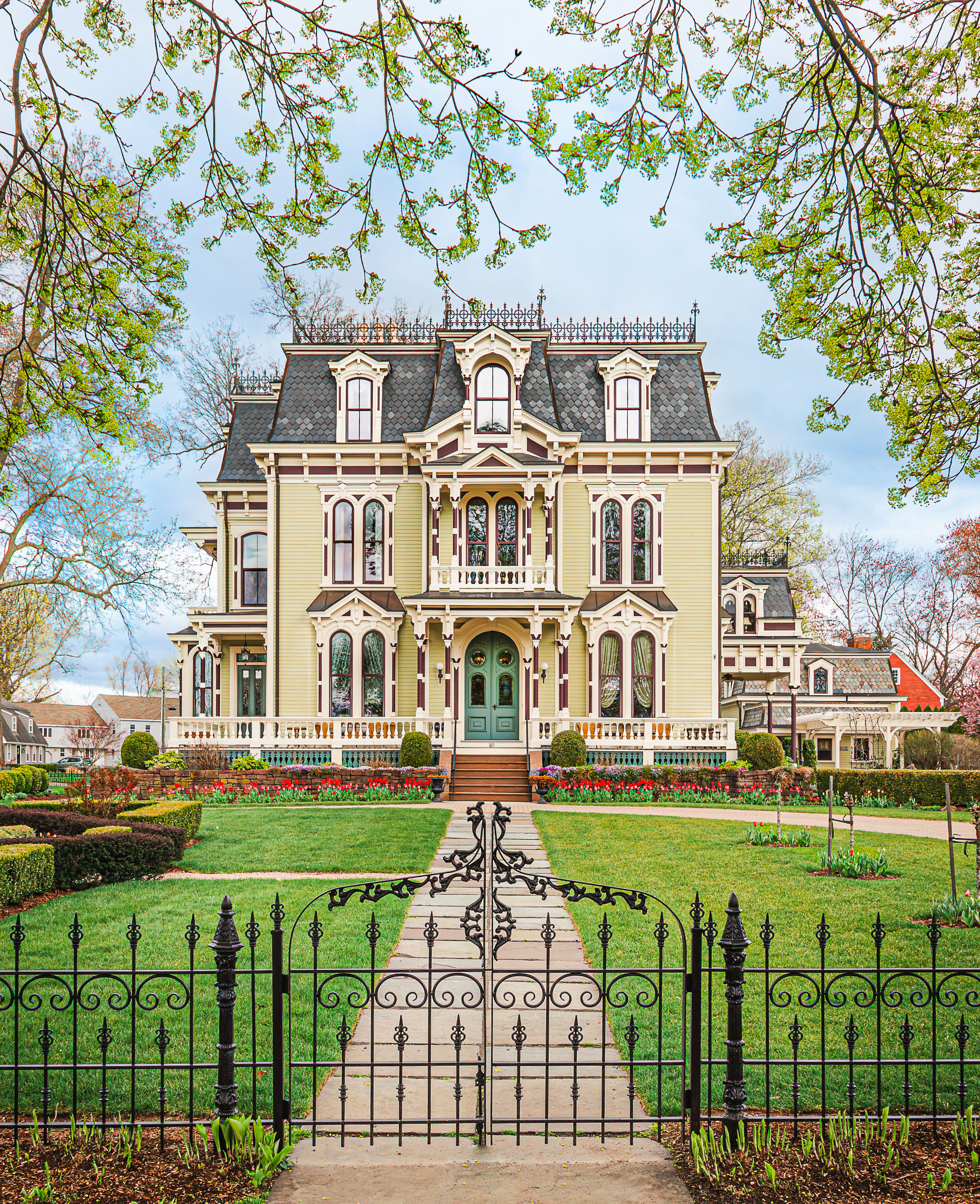 Historic Victorian-style home in Old Wethersfield, Connecticut, showcasing intricate architectural details, a mansard roof, colorful flower gardens, and decorative wrought-iron fencing.N4