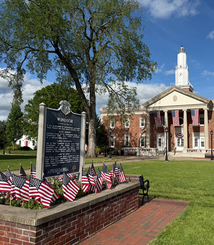 Historic Windsor Town Hall in Windsor, Connecticut, featuring its red brick facade, white columns, clock tower, American flags, and a historical marker overlooking the landscaped town green.