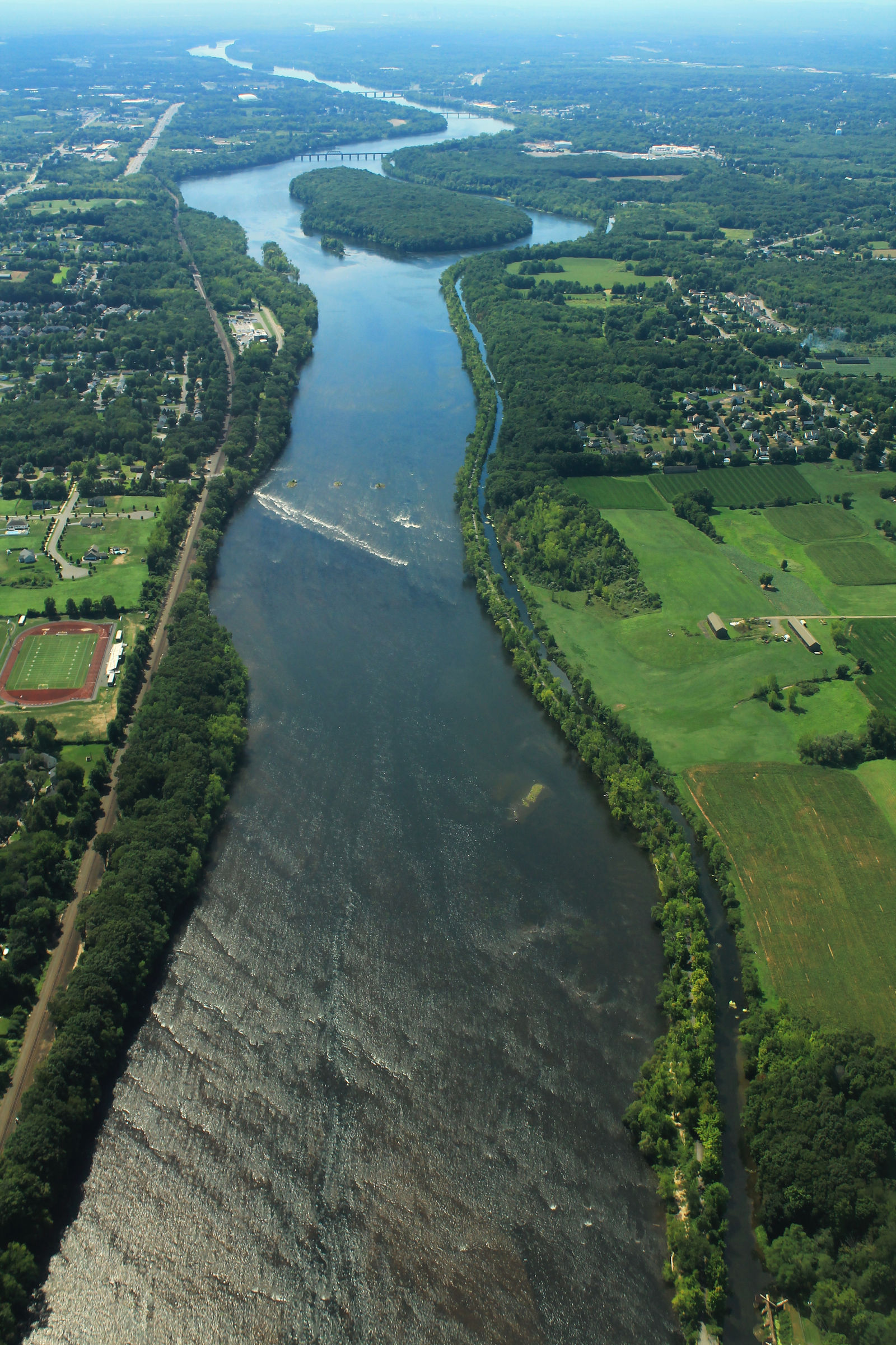Aerial view of the Connecticut River and surrounding Windsor Locks area, highlighting the river's winding waterways, wooded islands, lush green landscapes, and nearby residential neighborhoods.