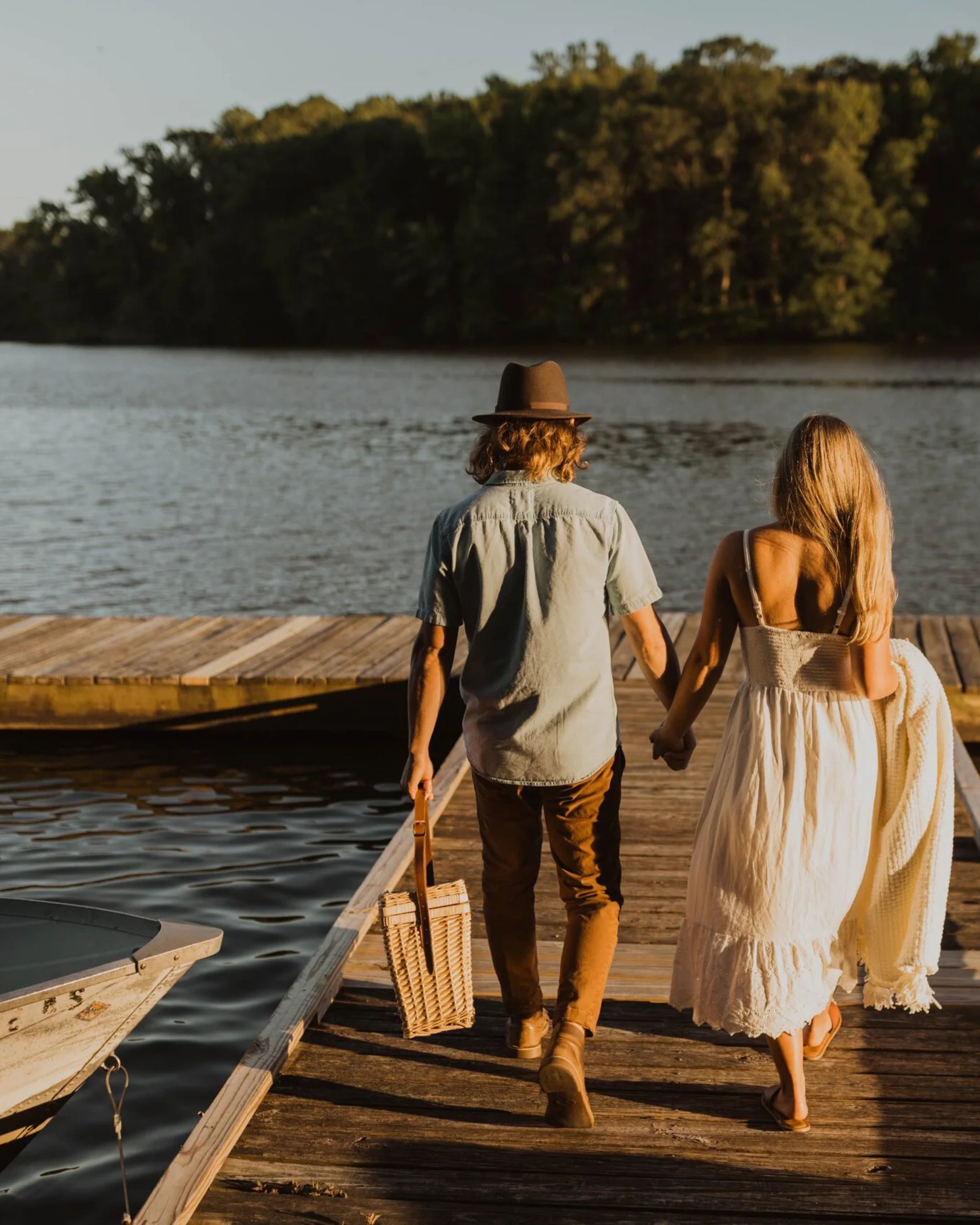 Couple walking hand in hand along a wooden dock at Lums Pond State Park in Bear, Delaware, during golden hour, with calm water, surrounding forest scenery, and a small boat resting beside the pier.