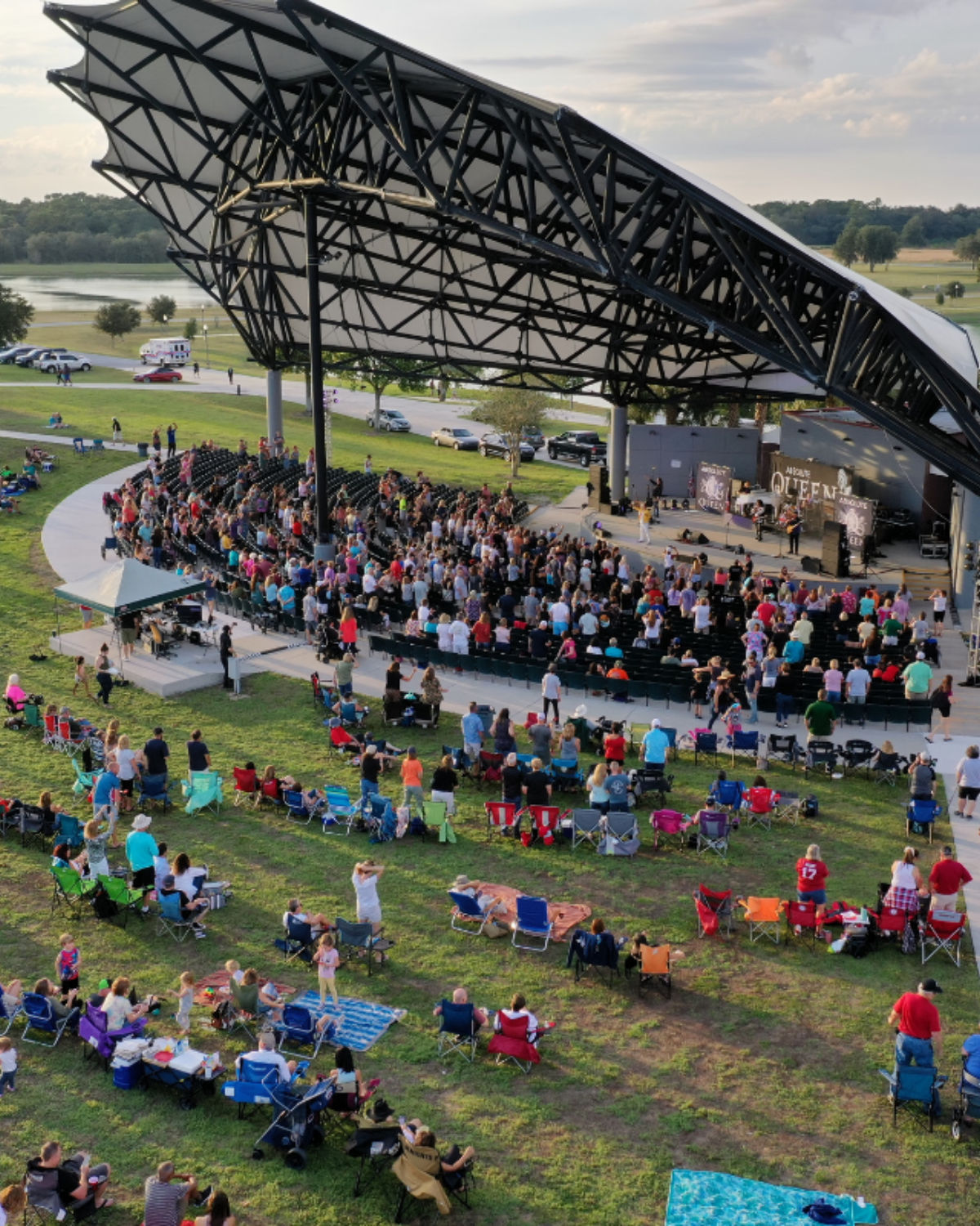 Crowd gathered for a concert at the Apopka Amphitheater in Apopka, Florida, with people seated on the lawn and under a large modern canopy covering the stage and main seating area.