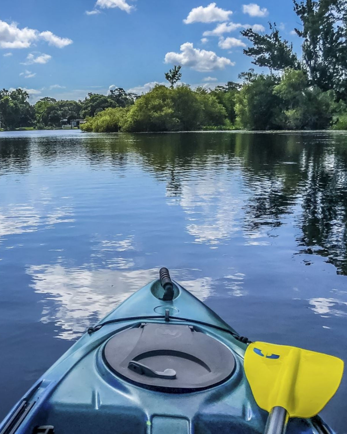Blue kayak with a yellow paddle floating on Triplet Lake in Casselberry, Florida, facing toward a tree-lined shoreline reflected in the calm water.