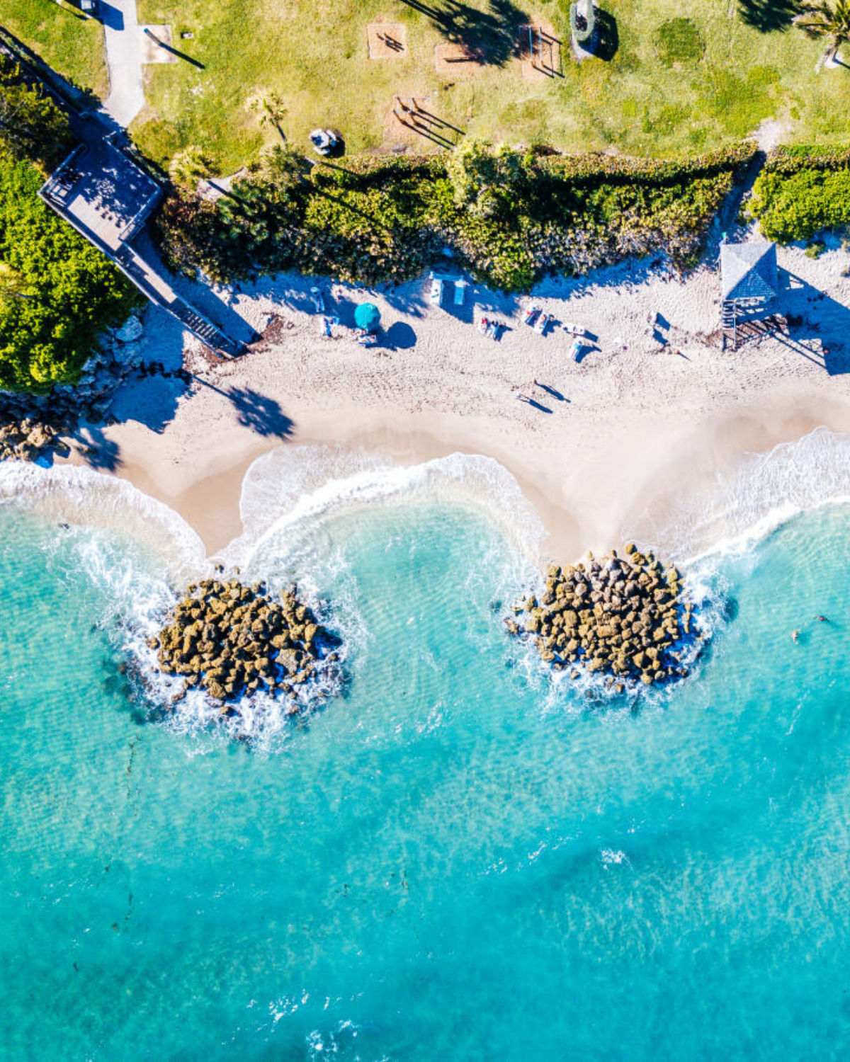 Overhead view of Deerfield Beach, Florida, featuring waves rolling onto a narrow strip of sandy shore lined with small rock formations, with sunbathers, beach umbrellas, and a grassy area shaded by palm trees