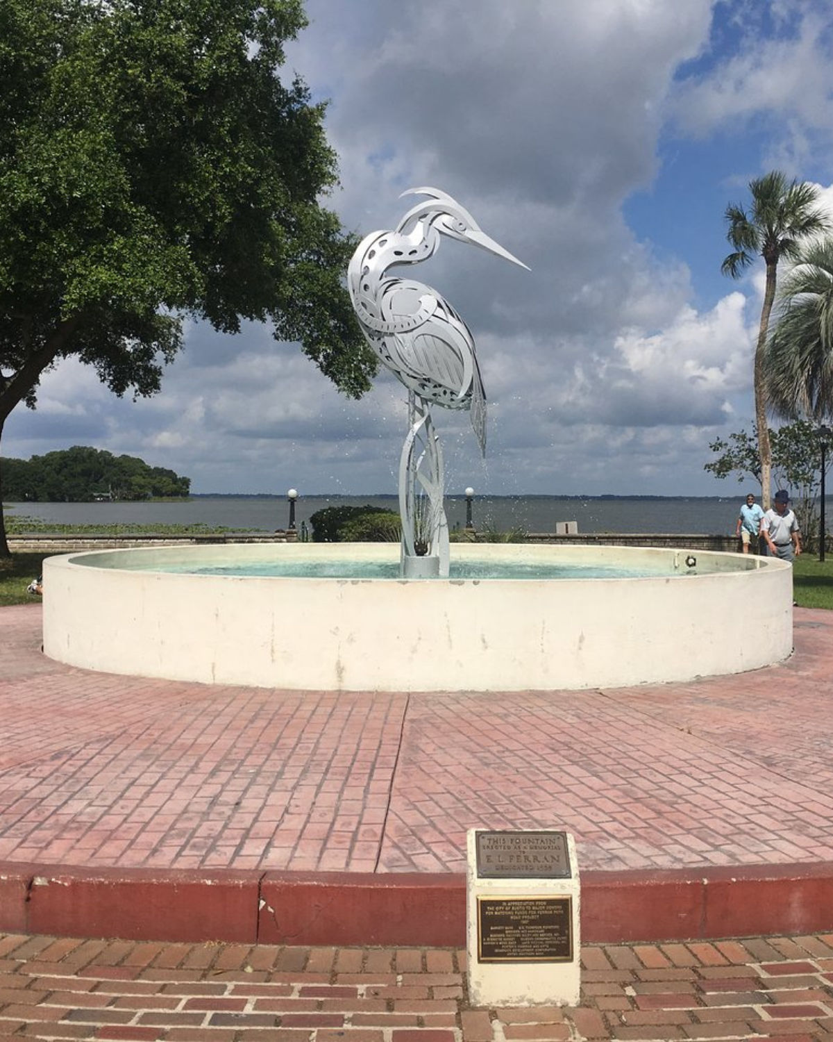 Metal heron fountain at Ferran Park in Eustis, Florida, with a sleek silver bird sculpture rising from a round pool on a brick plaza overlooking the lake.