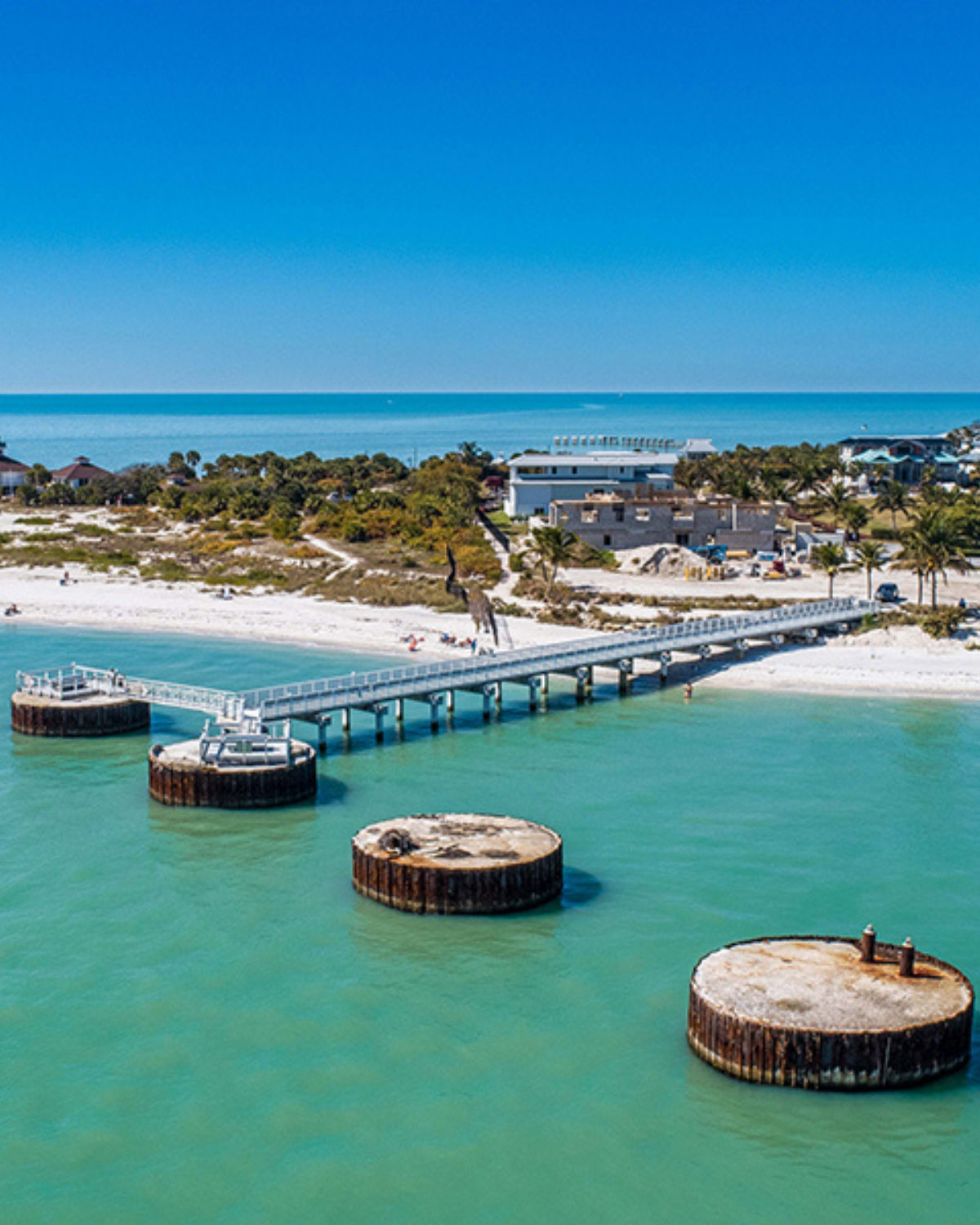 Aerial view of a white-sand beach and long pier at Fleming Island, Florida, stretching over clear turquoise water with round concrete pilings in the foreground and low coastal buildings and dunes behind.