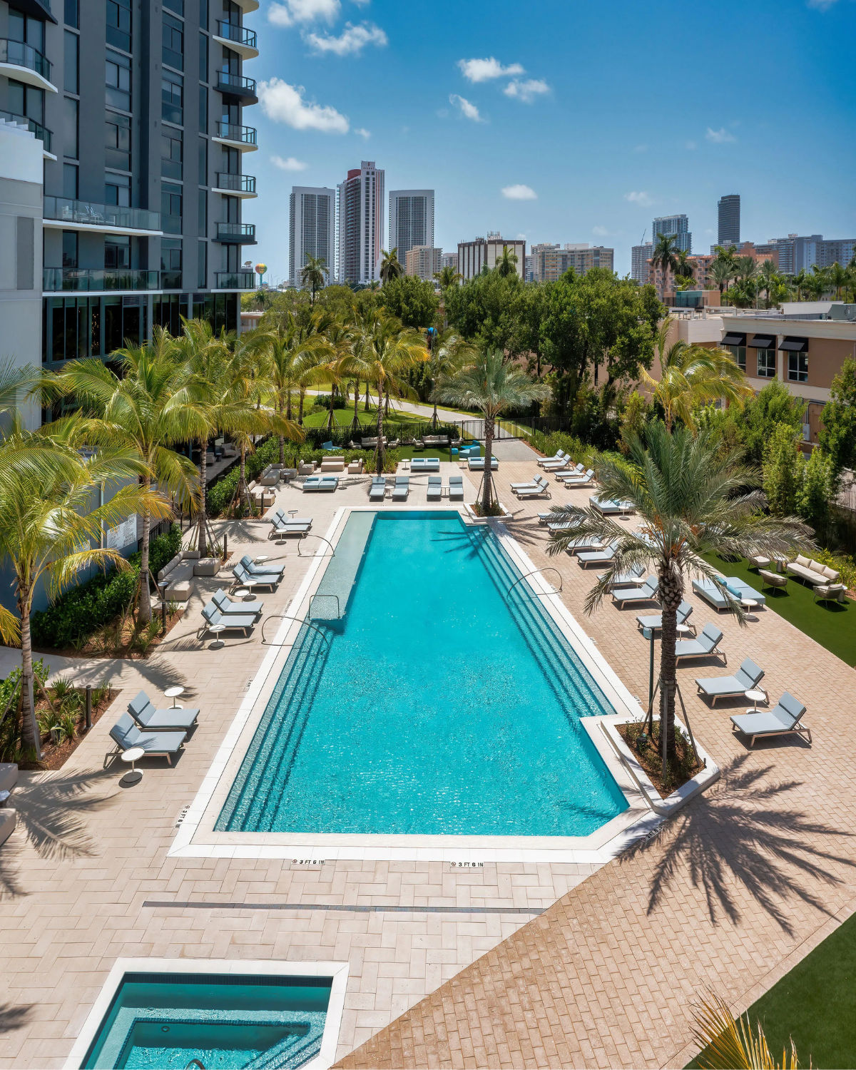 Resort-style pool deck in Hallandale Beach, Florida, with a long rectangular swimming pool lined by lounge chairs and palm trees, set below modern high-rise condos and city towers in the background.