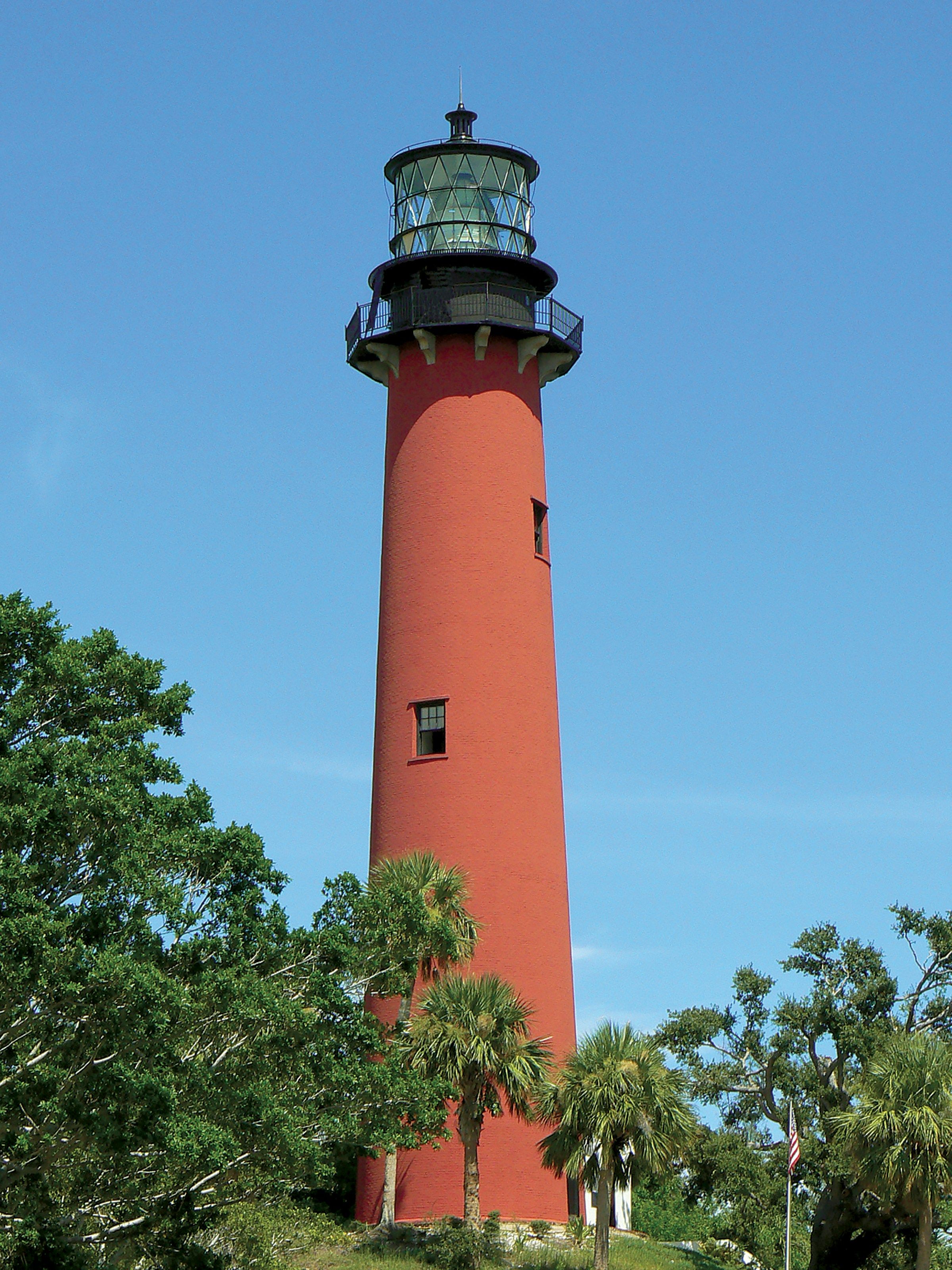 Iconic red Jupiter Inlet Lighthouse rising above lush green tropical trees against a bright blue sky