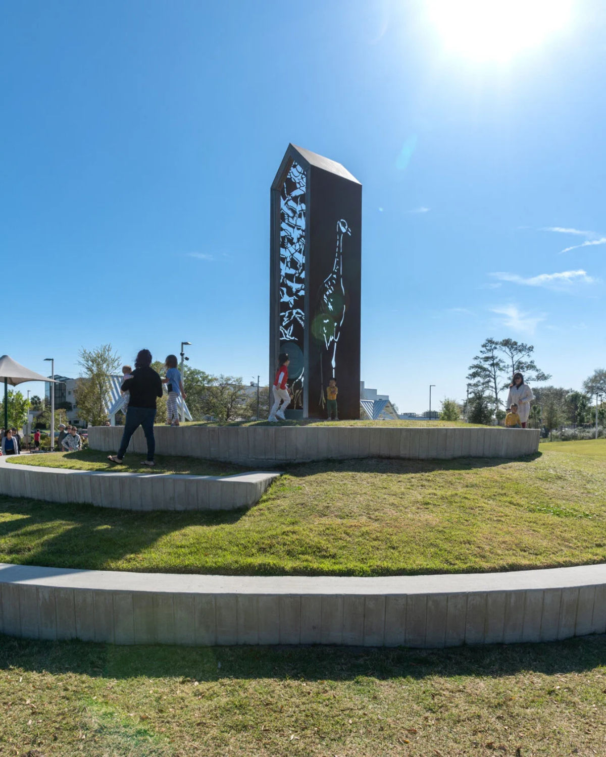 Families walking around a tiered grassy mound at Reiter Park in Longwood, Florida, centered on a tall metal sculpture tower with cut-out bird and heron designs.