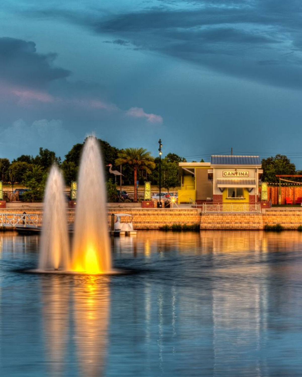 Colorful lakeside park in Oviedo, FL featuring decorative fountains, a pavilion, and lush tropical landscaping at dusk