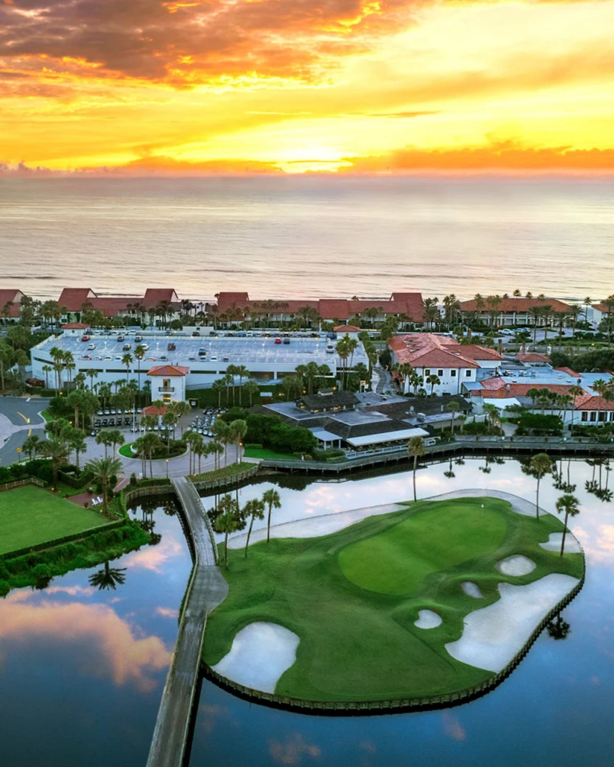 Aerial view of Ponte Vedra Inn’s island green in Ponte Vedra Beach, Florida, with a small putting green ringed by sand bunkers in a reflective lagoon, connected to the resort by a narrow bridge and set against an ocean sunrise.