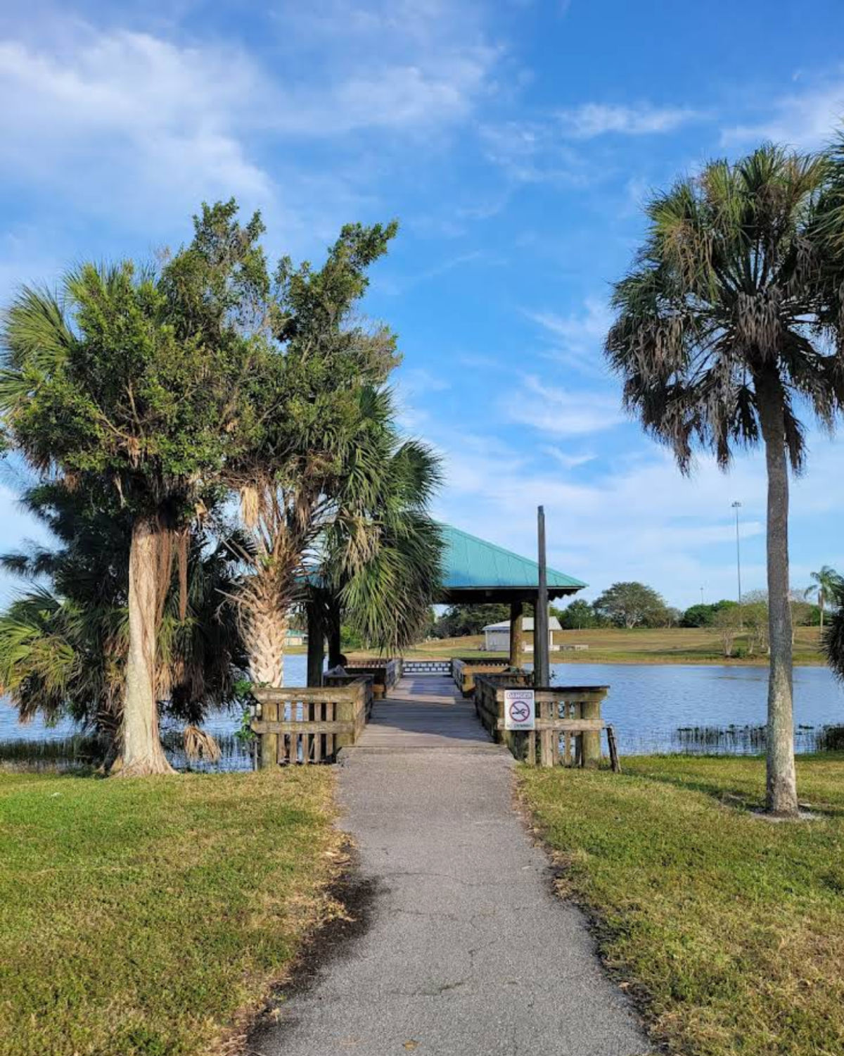 Walkway leading to wooden fishing pier and small covered pavilion on the lake at C.B. Smith Park in Pembroke Pines, Florida, framed by tall palm trees