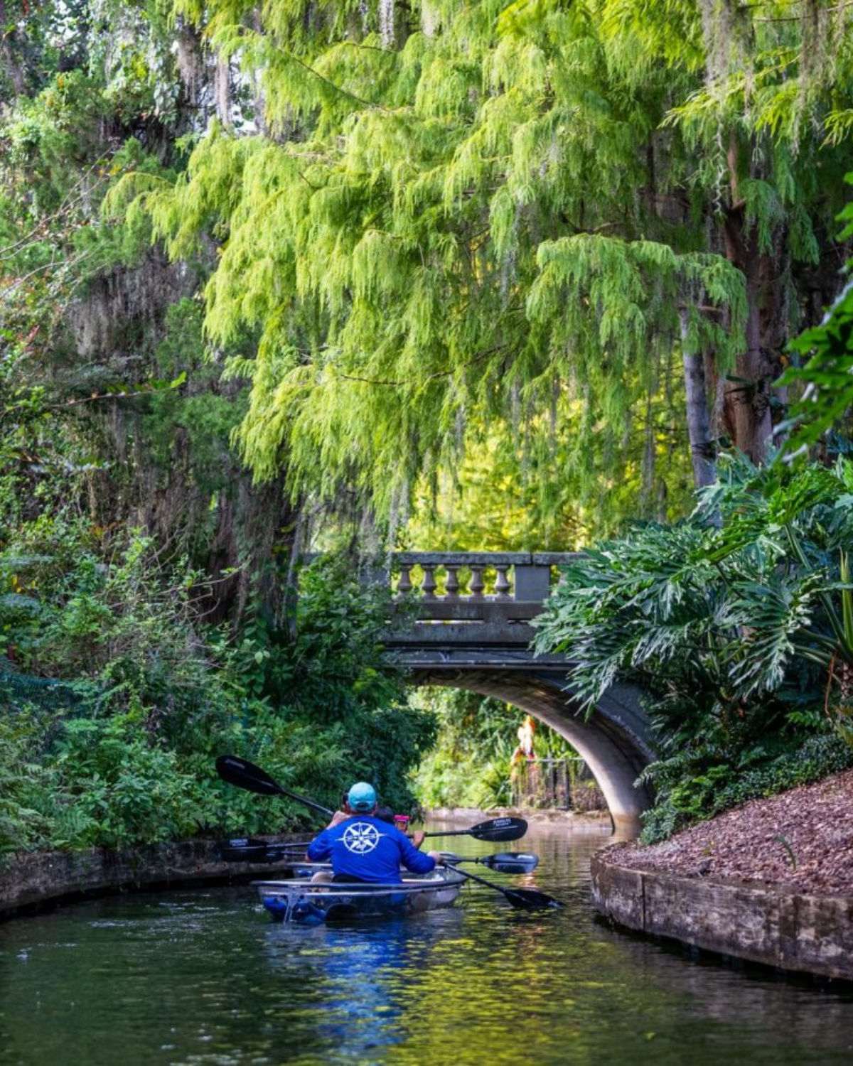 Kayakers paddling through a narrow, tree-lined canal on the Winter Park Chain of Lakes in Winter Park, Florida, passing under a small arched bridge between the residential lakes.
