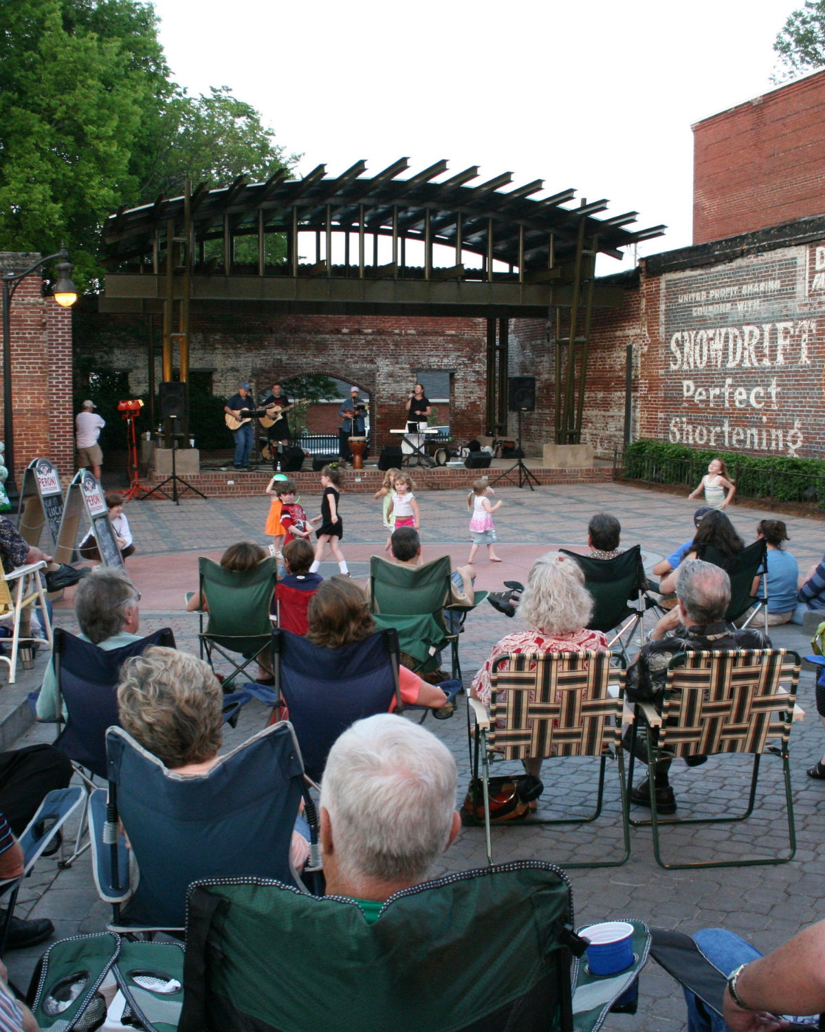 Evening community concert at OŐNeal Plaza in downtown Douglasville, Georgia, with families seated in lawn chairs watching a live band perform on a brick stage under a modern metal canopy while children dance on the plaza