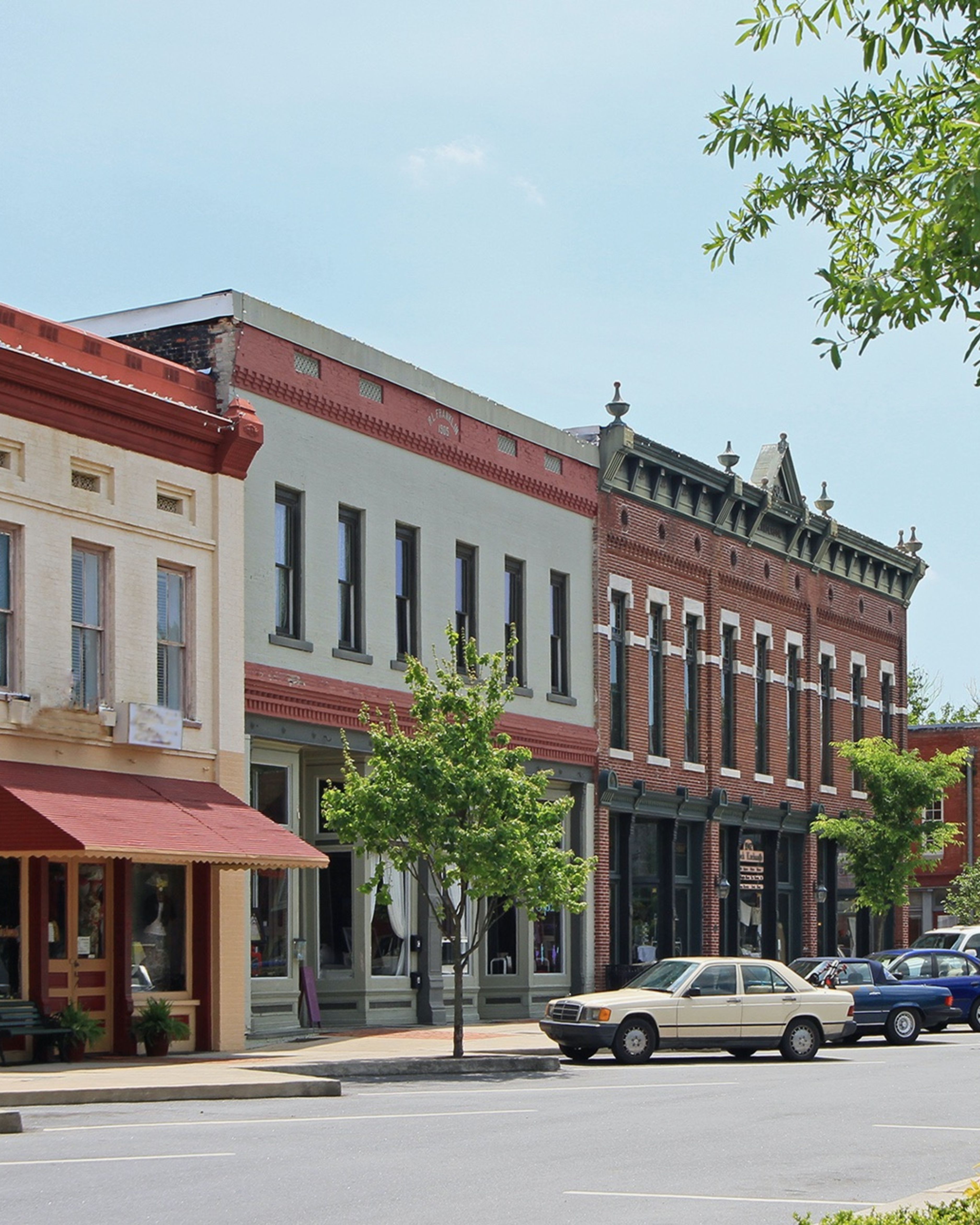 Historic downtown Gainesville, Georgia streetscape featuring brick storefronts, local businesses, parked cars, and classic Southern architecture lining a sunny small-town commercial district.